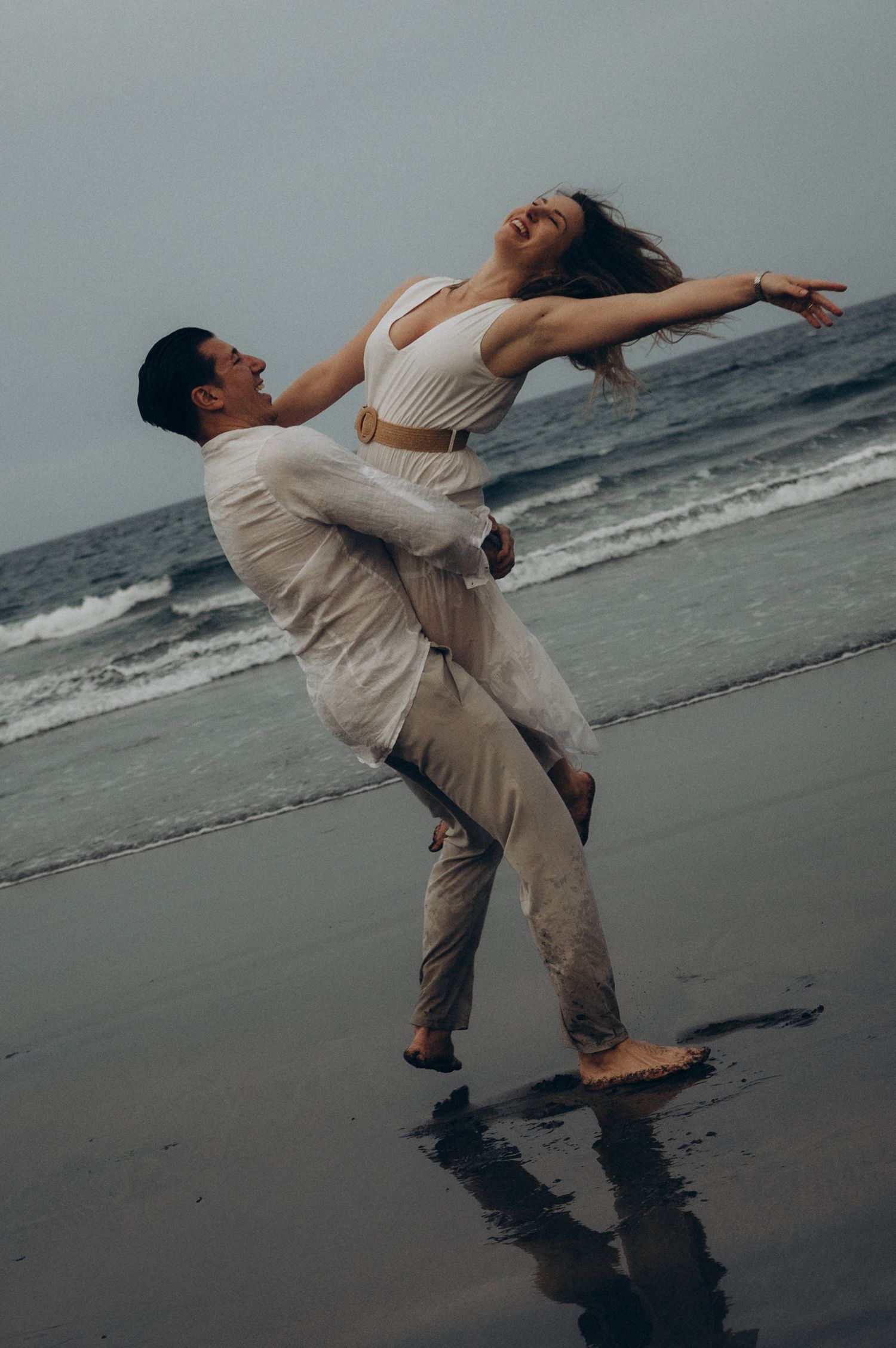 Couple dancing joyfully on a beach, woman in white crop top leaning back with arms spread, man lifting her at the shoreline.