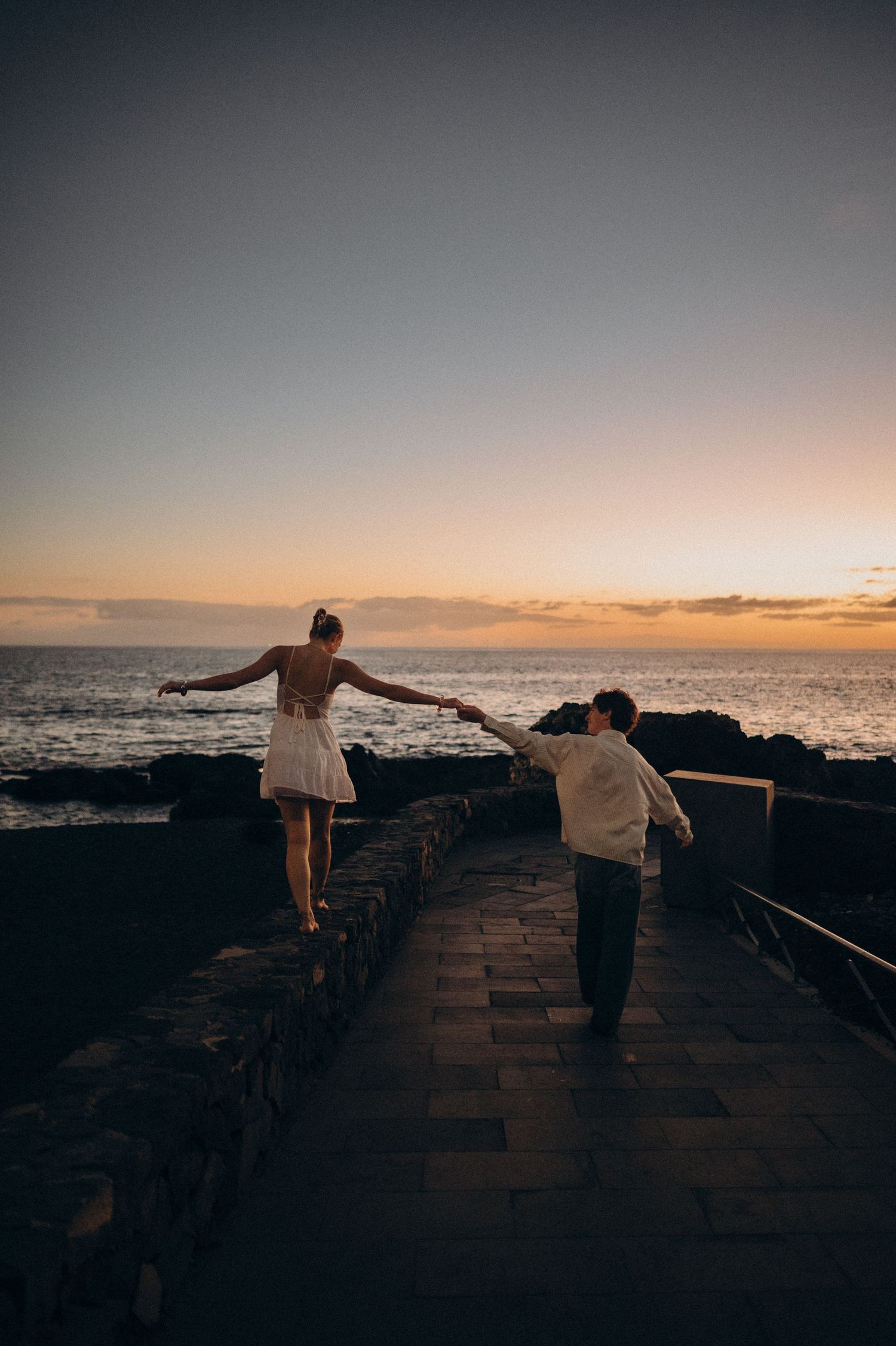Couple holding hands at sunset on a coastal stone walkway, woman in white dress balancing on wall by the ocean.