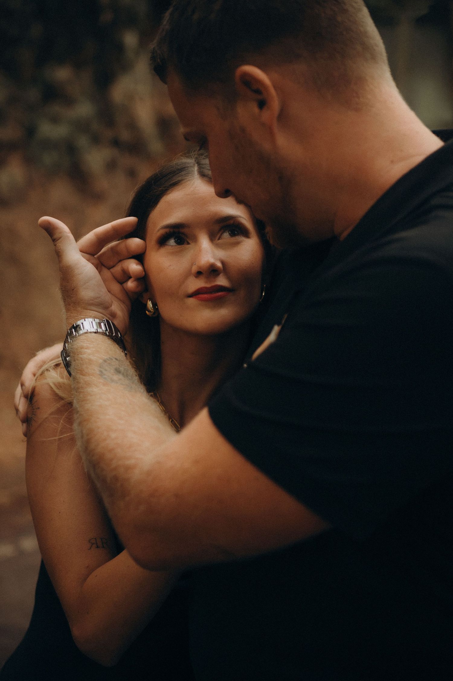A couple embraces tenderly, the woman gazing up lovingly at the man in a black t-shirt wearing a watch.
