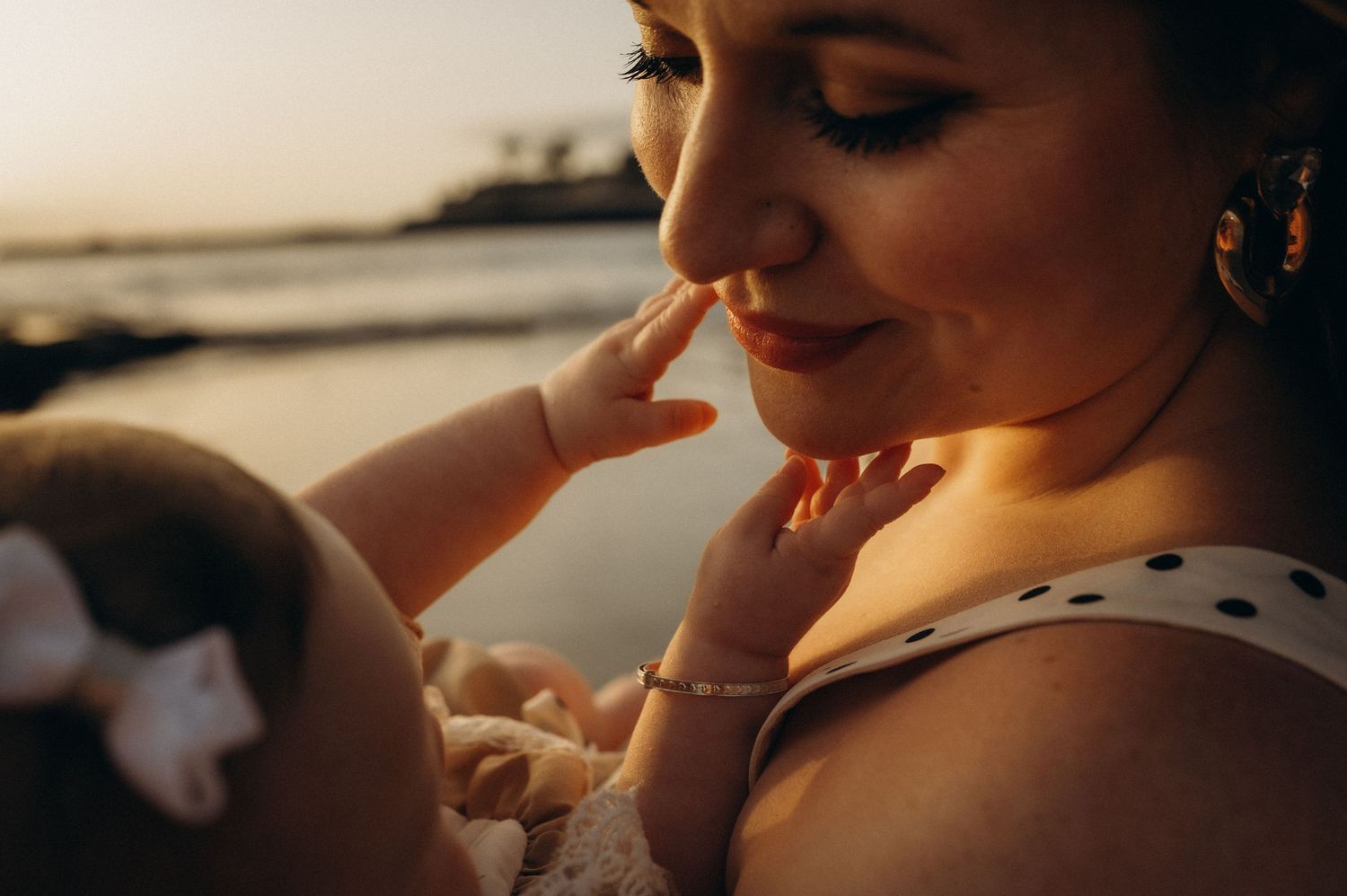 A mother tenderly holds her newborn baby at golden hour near the water, bathed in warm sunset light. Costa Adeje, Tenerife