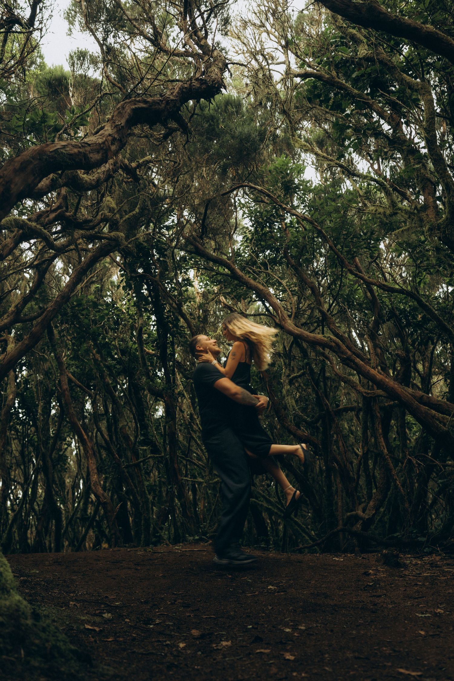 Couple embracing in a dark mystical forest, man lifting woman as trees twist overhead in moody woodland setting.
