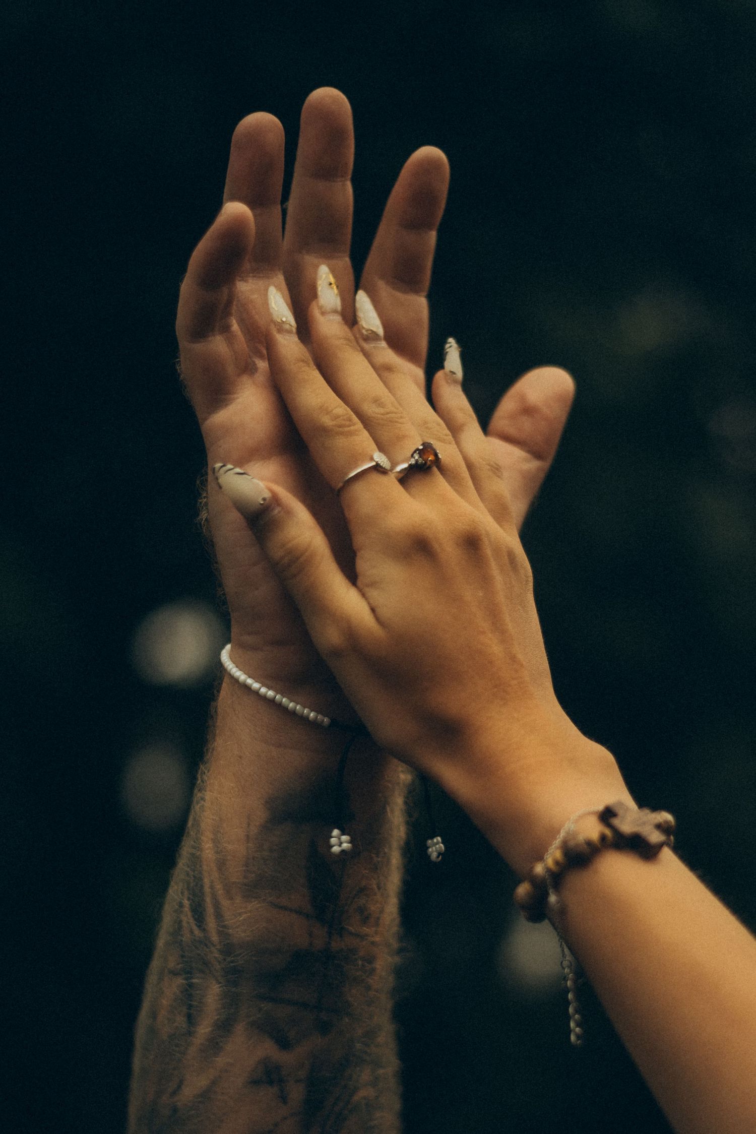 Three hands raised together, adorned with rings, bracelets, and tattoos, against a dark background.