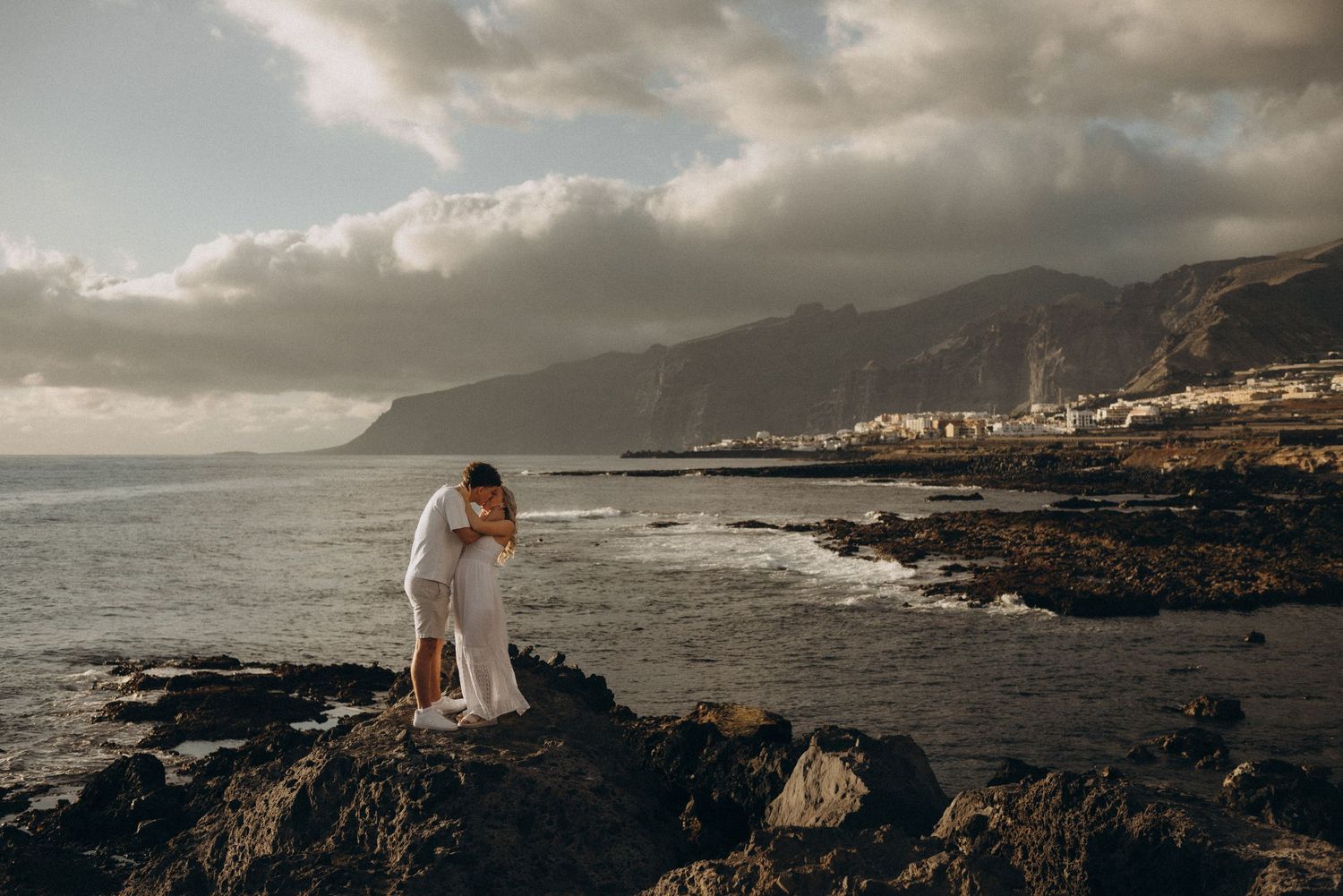 Couple embracing on rocky coastal cliffs with dramatic ocean waves and mountain backdrop during golden hour.
