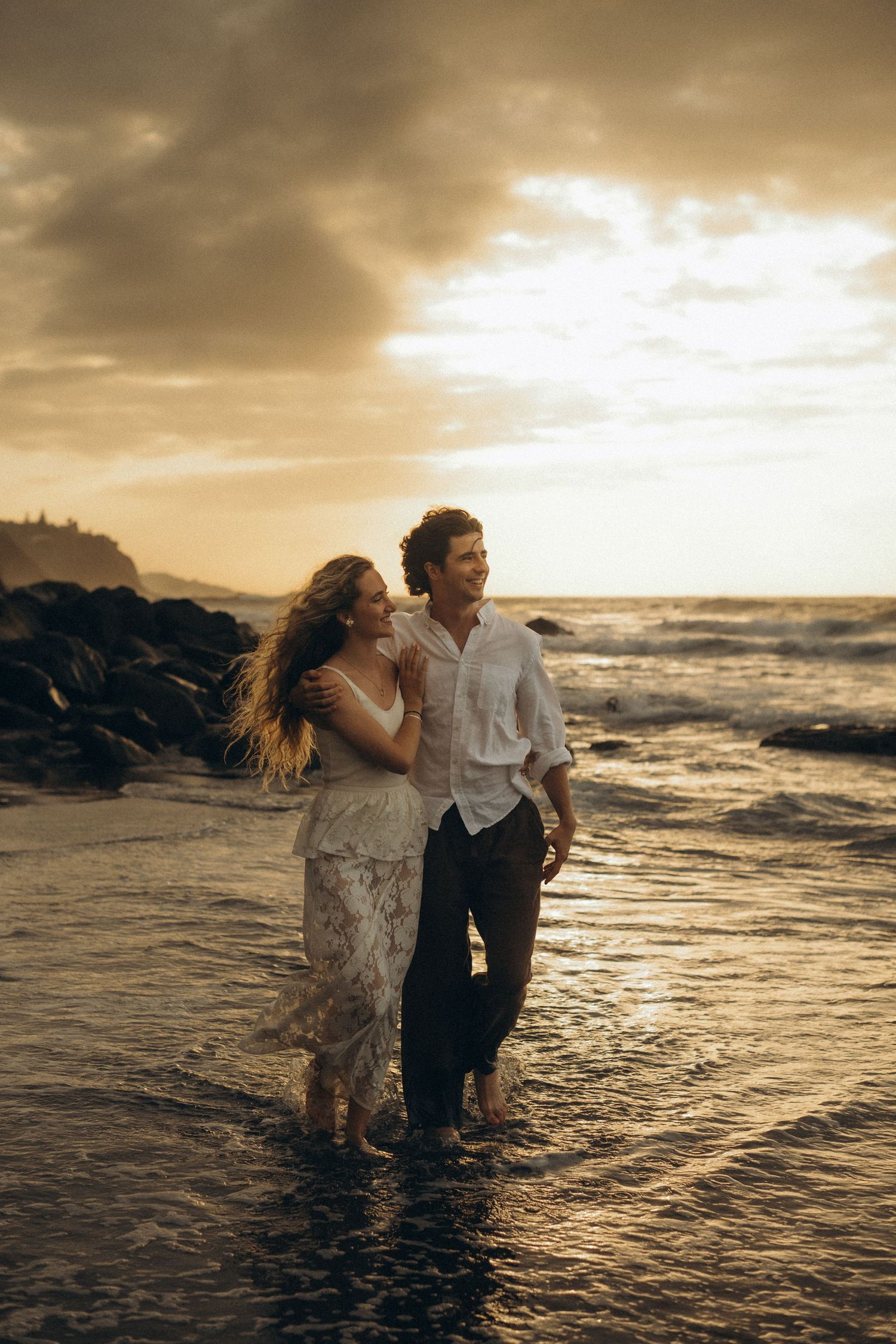 Couple walking barefoot on a rocky beach at golden sunset, woman in lace dress, man in white shirt.