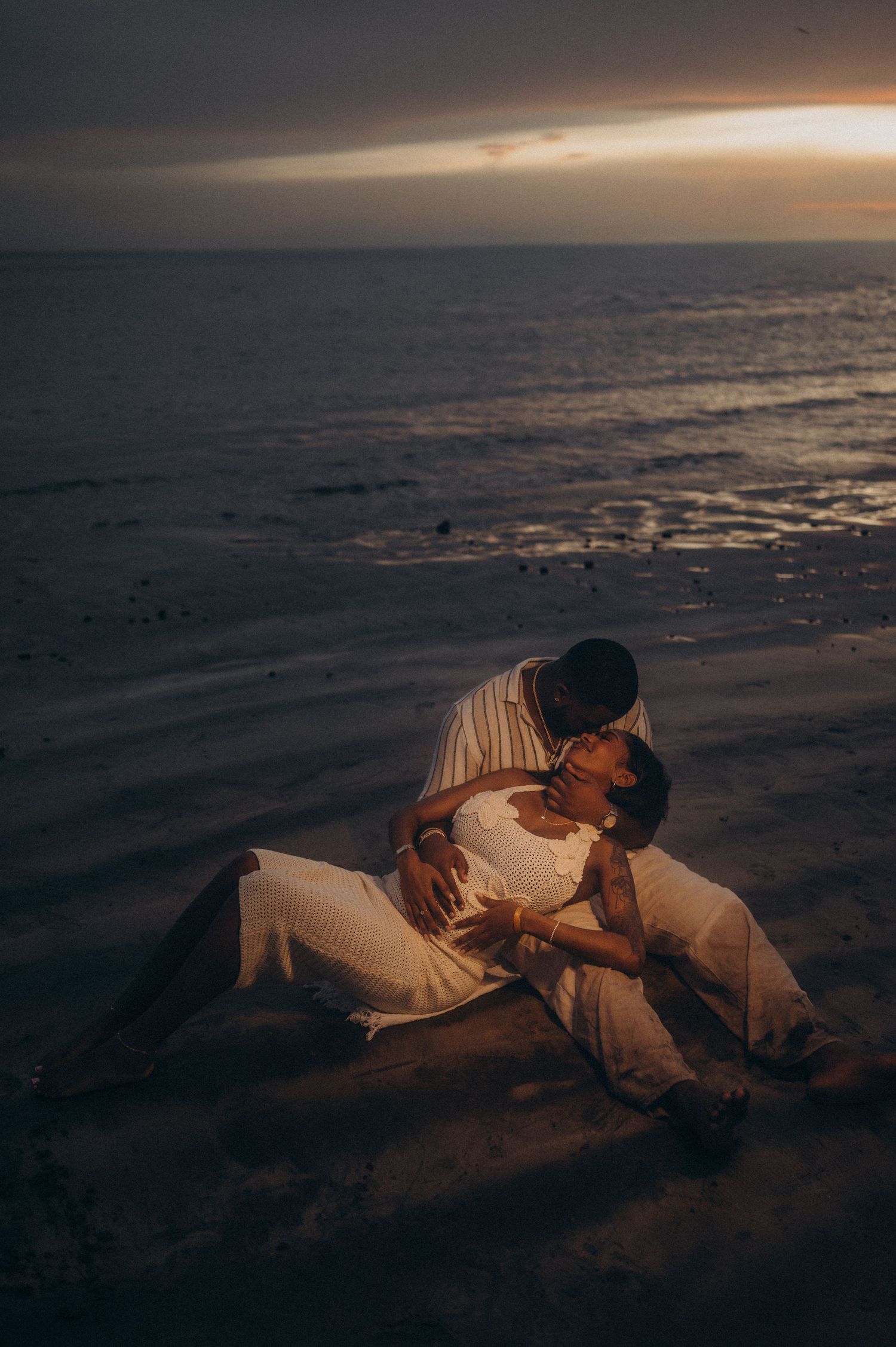 Pregnant woman lying in husband’s arms while he caresses her belly at sunset, Playa Fañabé