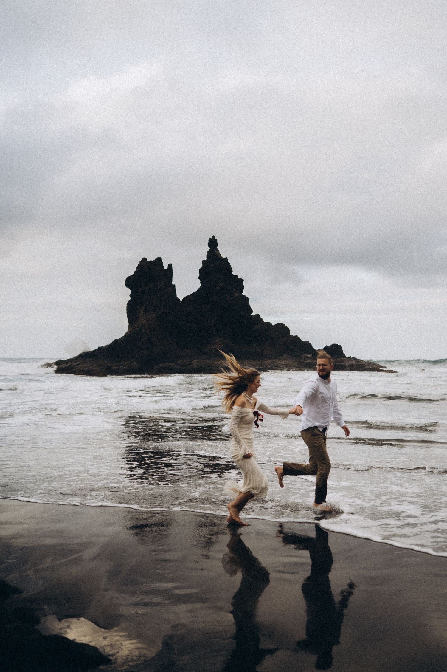 Couple running on Playa de Benijo beach in Tenerife during photoshoot with dramatic rock formations and moody skies.