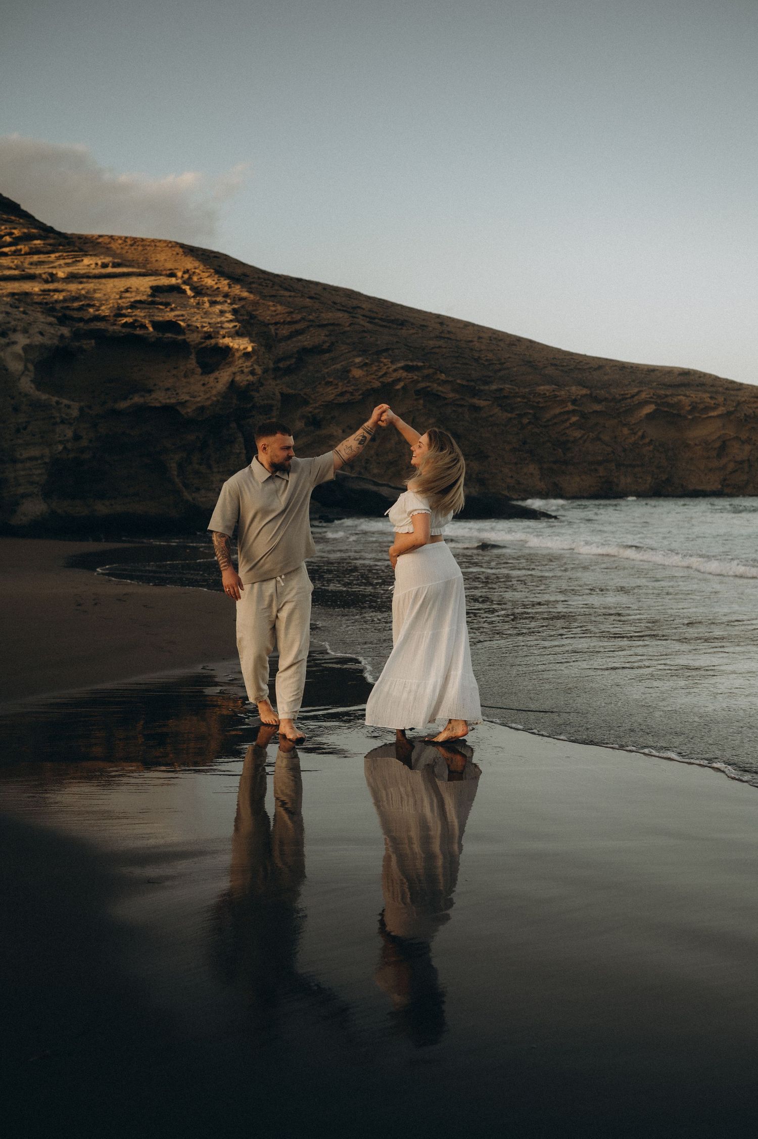 Couple dancing on a black sand beach at sunset, with rocky cliffs and ocean waves reflecting in the wet sand.