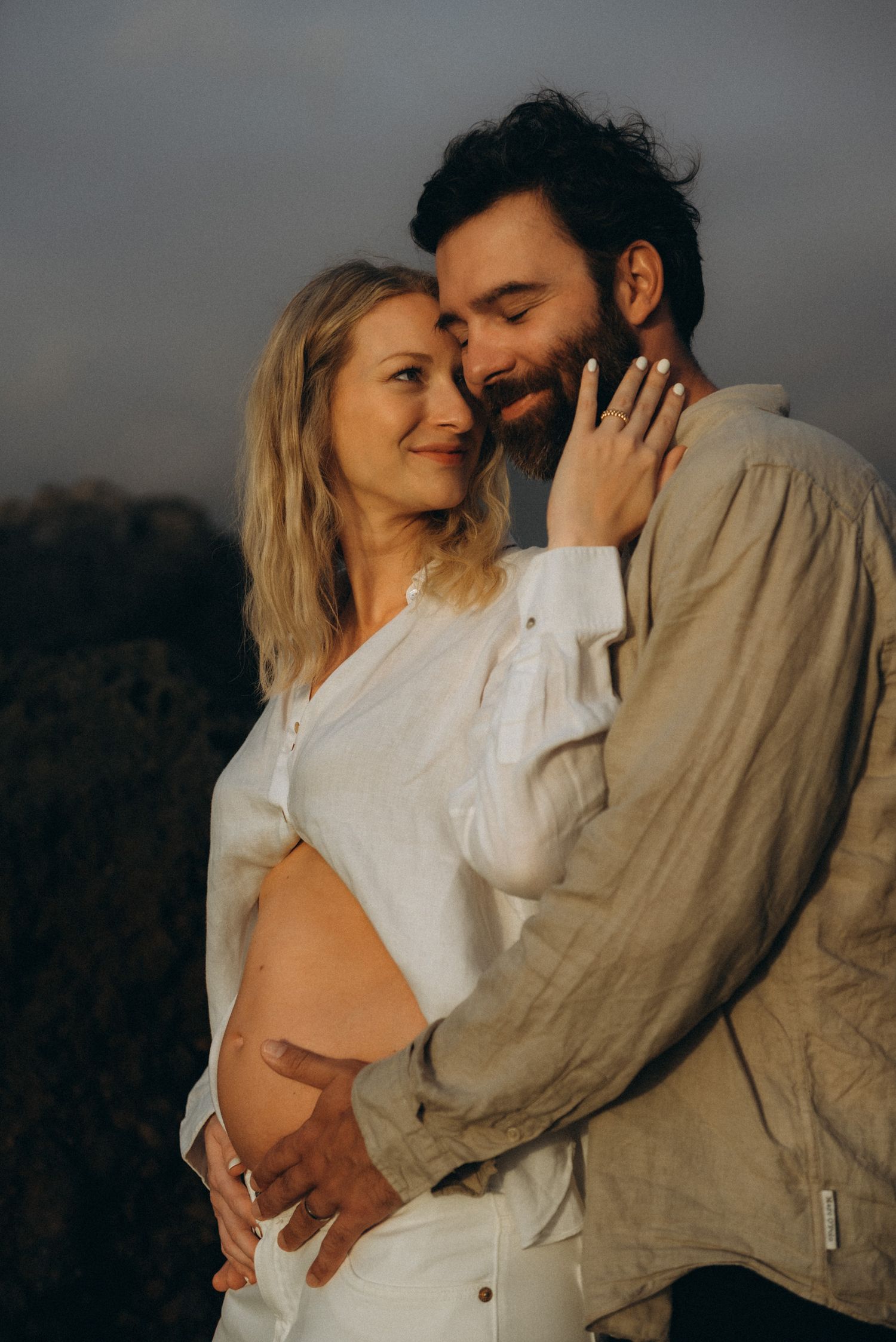 Couple embracing outdoors at sunset, pregnant woman in white shirt with partner holding her baby bump.