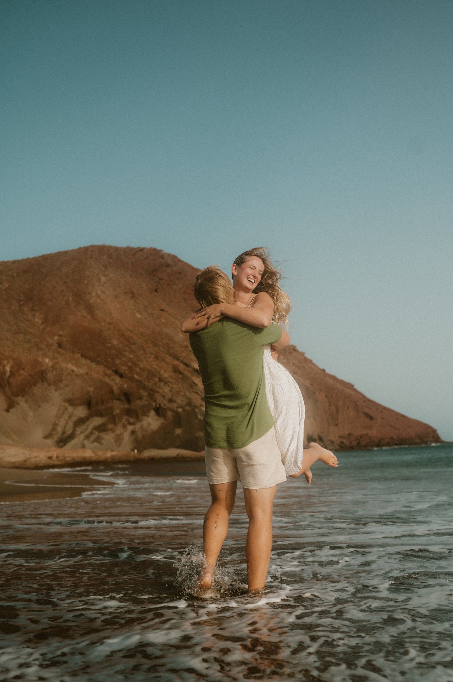 Young couple on Playa Tejita, he holds her in his arms from a different angle, she leans back with hair flowing in the wind, Montaña Roja vi
