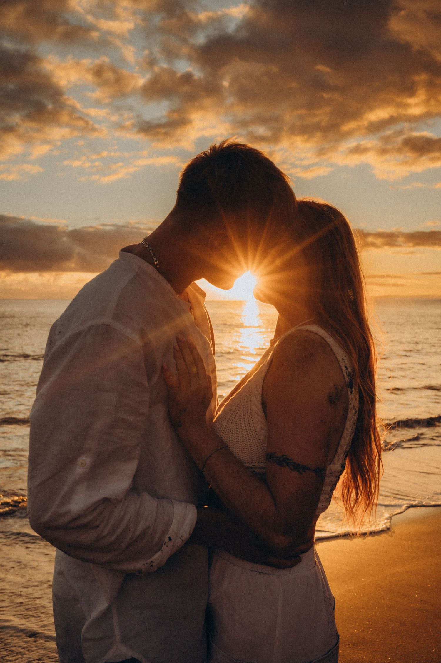 Silhouetted couple embracing during sunset photoshoot on Tenerife beach with dramatic orange clouds and ocean waves.