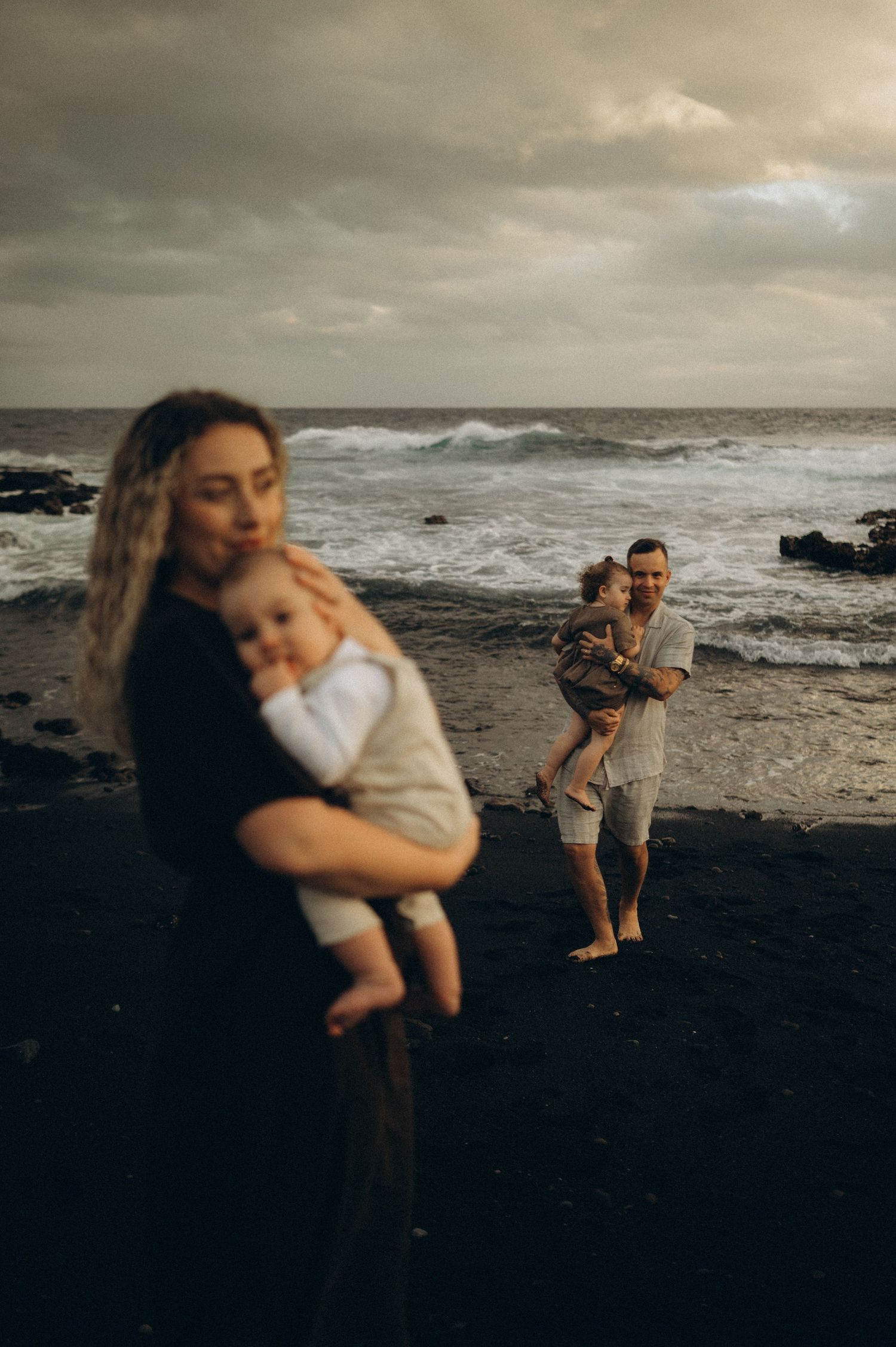Family of four on a dark sandy beach, mother holding baby in foreground, father carrying toddler by moody ocean waves. Playa La Jaquita