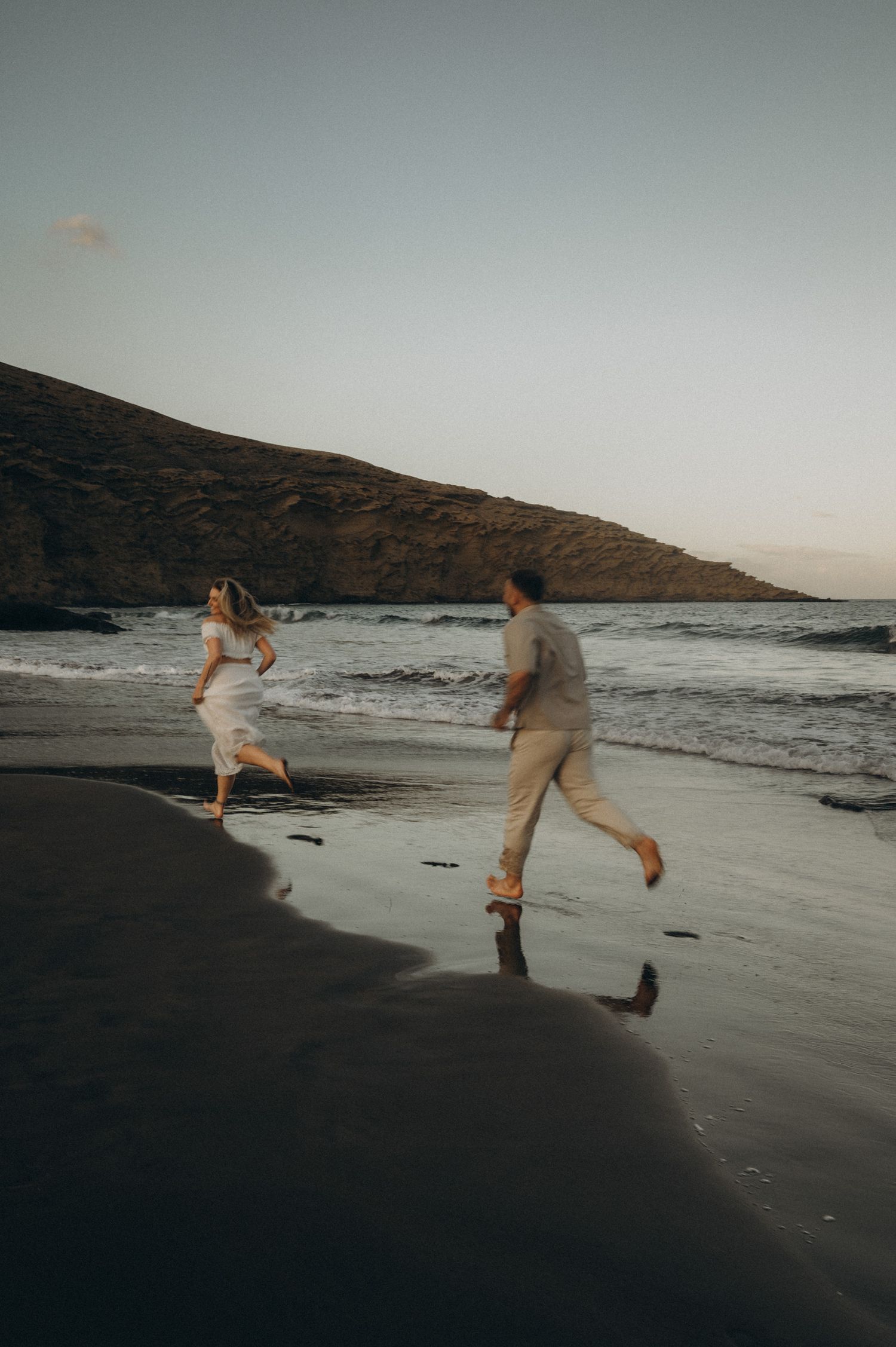 Couple running barefoot on a dark sandy beach at dusk with ocean waves and a rocky hill in the background.