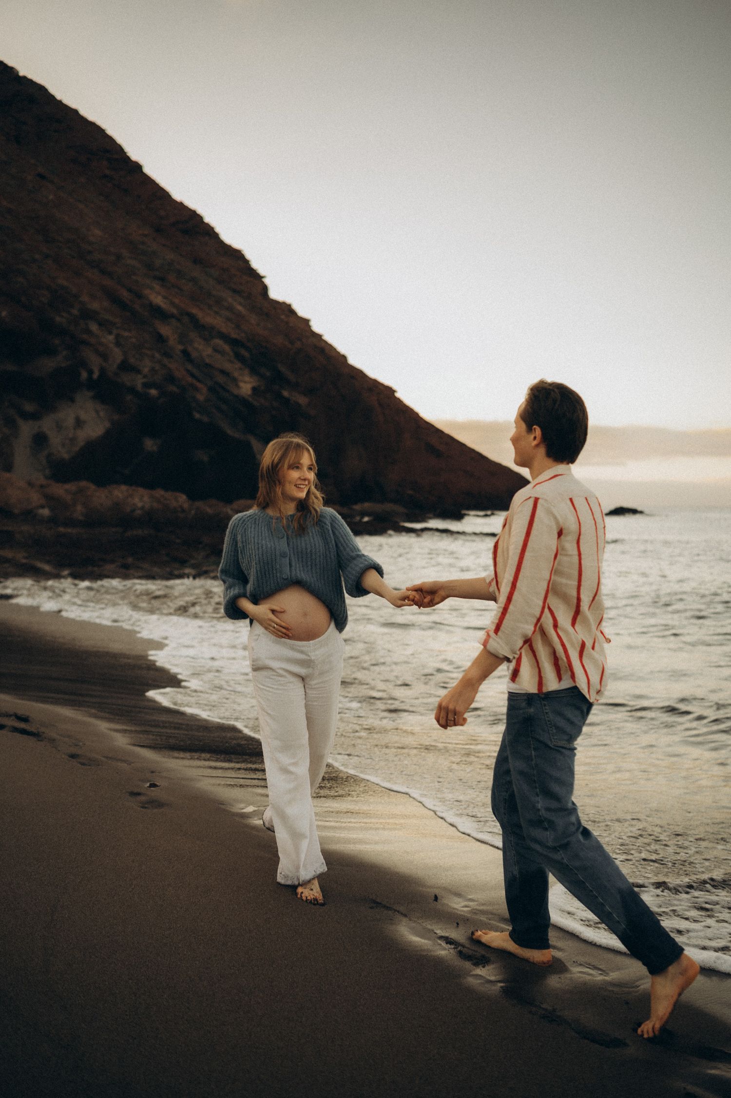 Pregnant woman and partner holding hands walking barefoot on a beach at sunset near rocky cliffs.