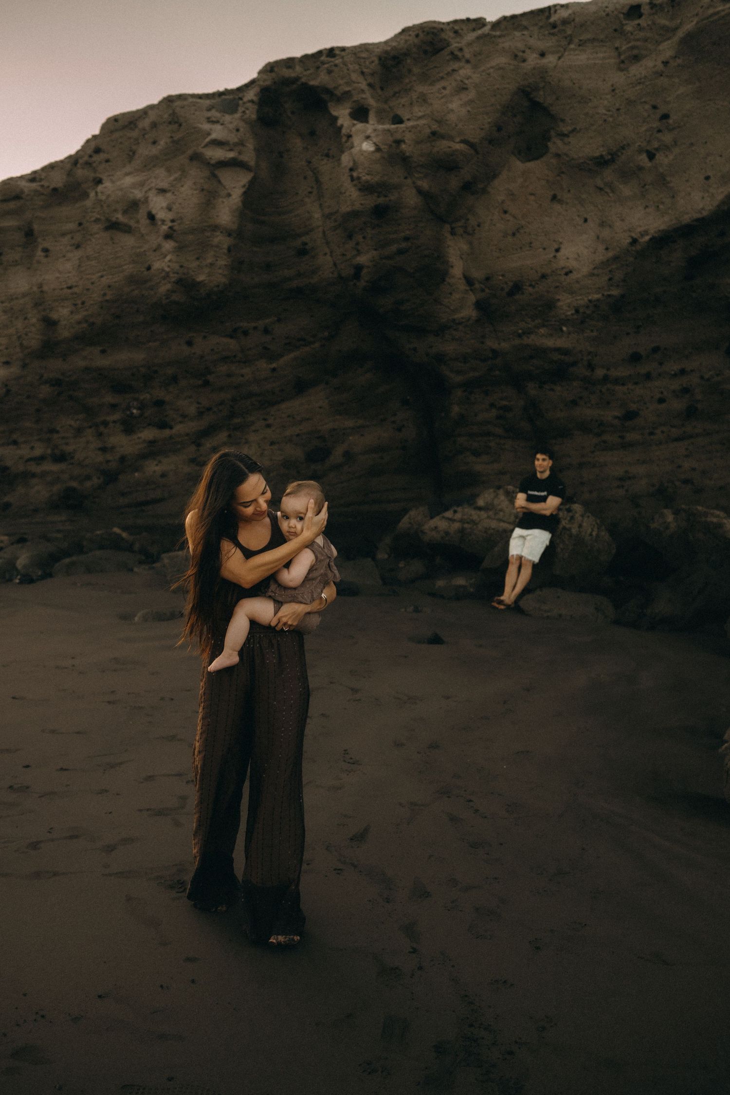 A mother holds her baby on a dark sandy beach with rocky cliffs, while a man stands in the background.