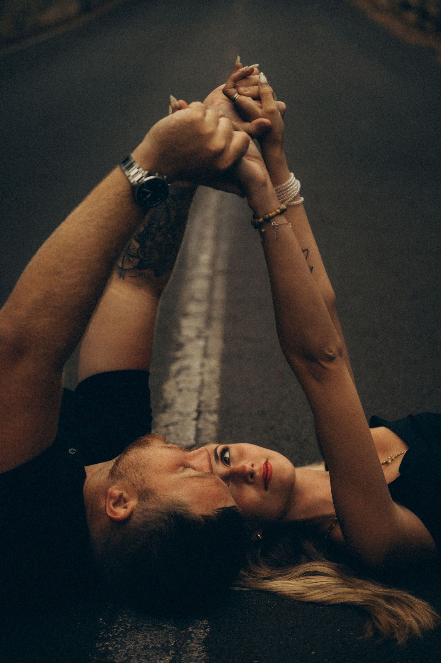 Couple lying on road holding hands up, man with tattoos and watch, woman with bracelets, romantic dark aesthetic.