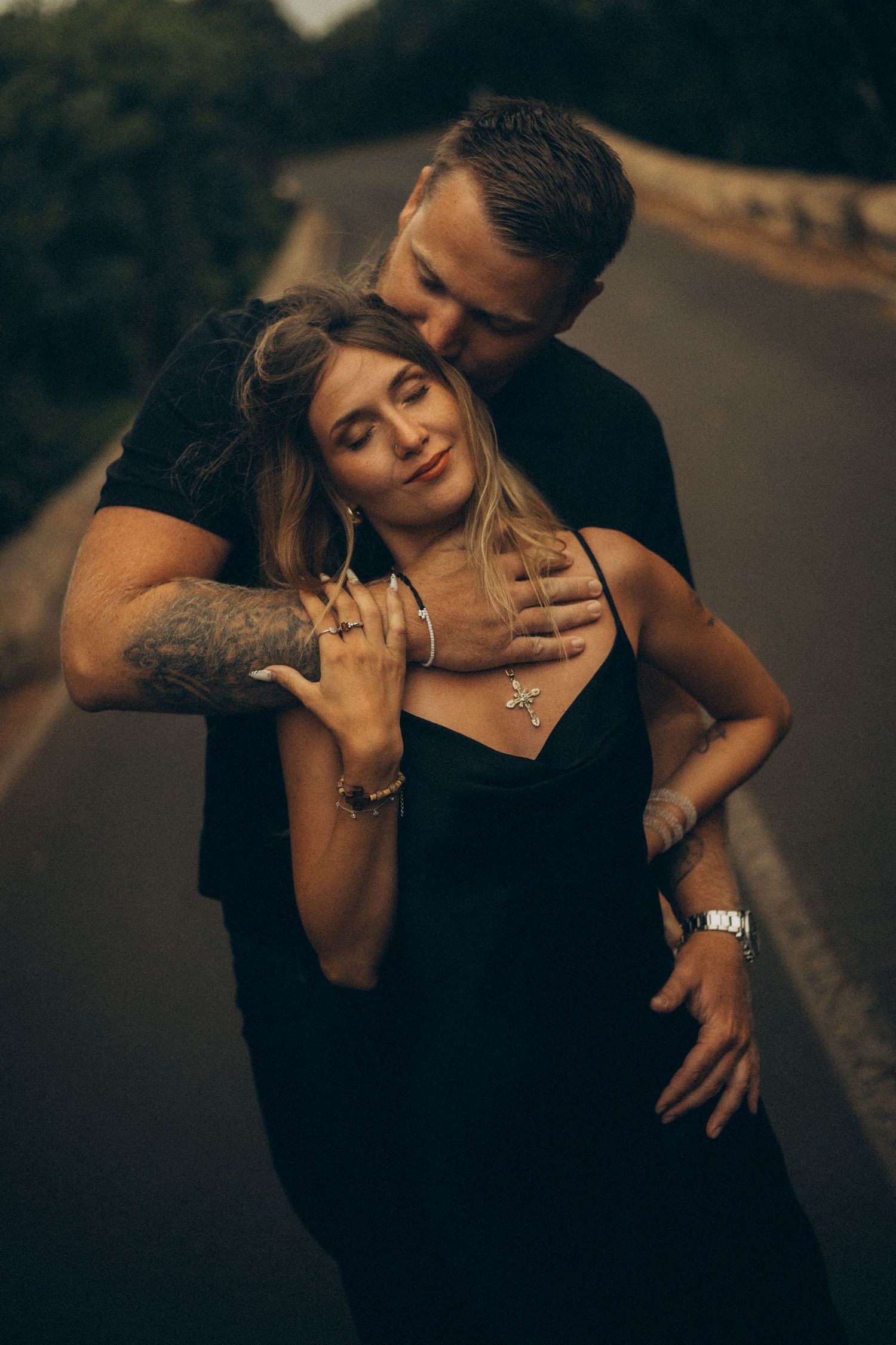 Romantic couple in black outfits embracing on a road, man kissing woman's head, both wearing watches and jewelry.