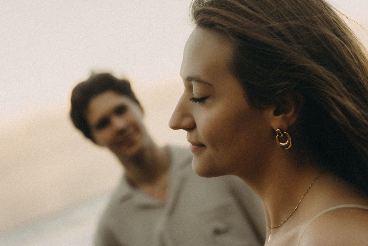 A woman with hoop earrings smiles softly in the foreground while a man in a polo shirt looks on in the blurred background.