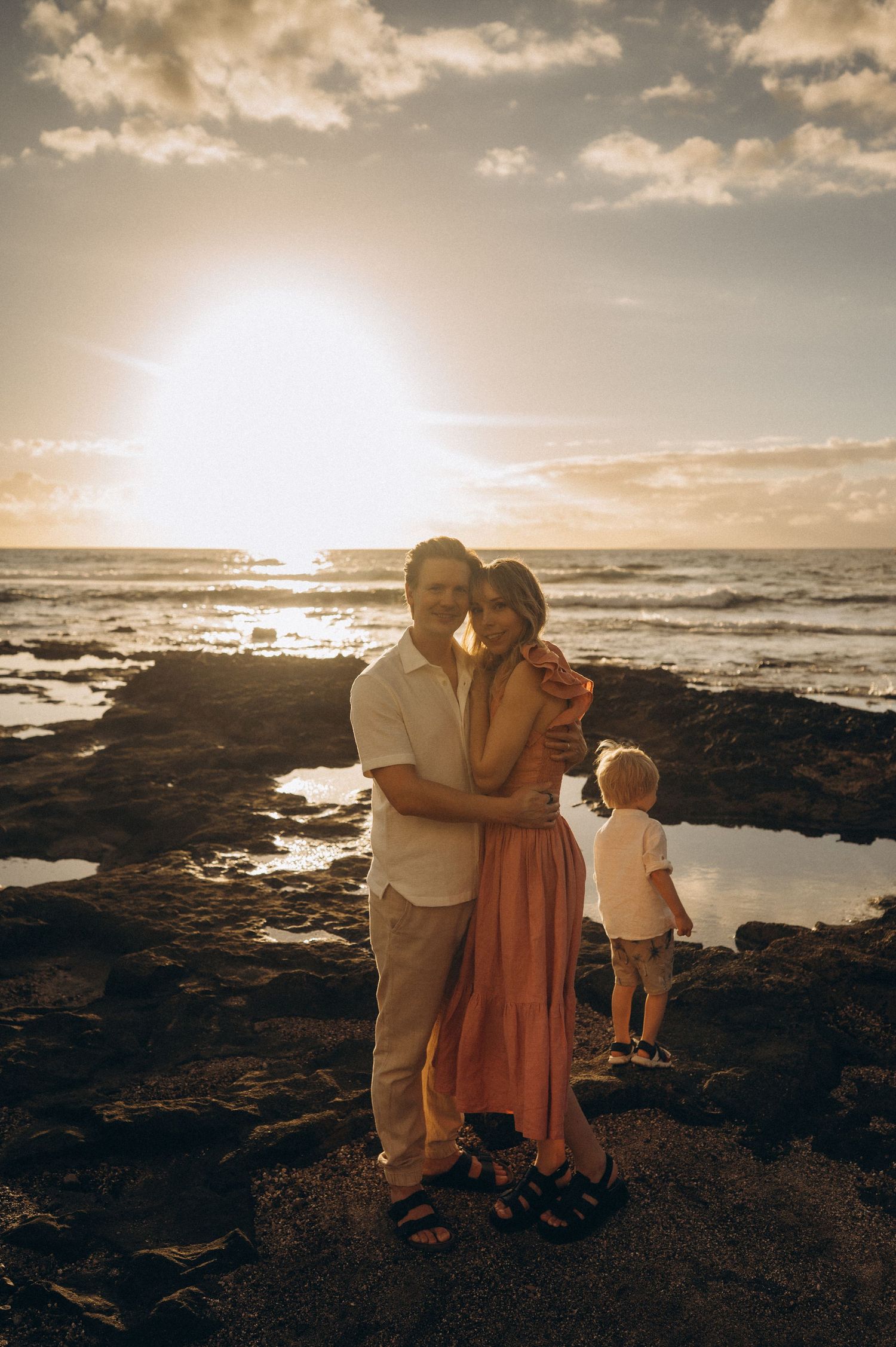 Family on beach at sunset, parents holding each other while child plays near sea, Las Americas