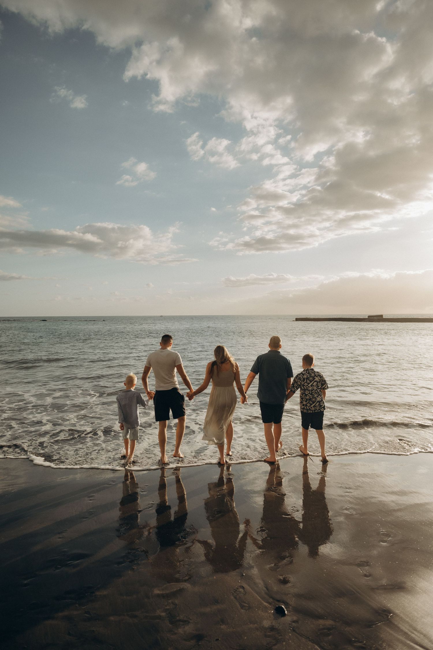 Parents and three sons standing hand in hand watching sea with mirrored sand, Playa Fañabé