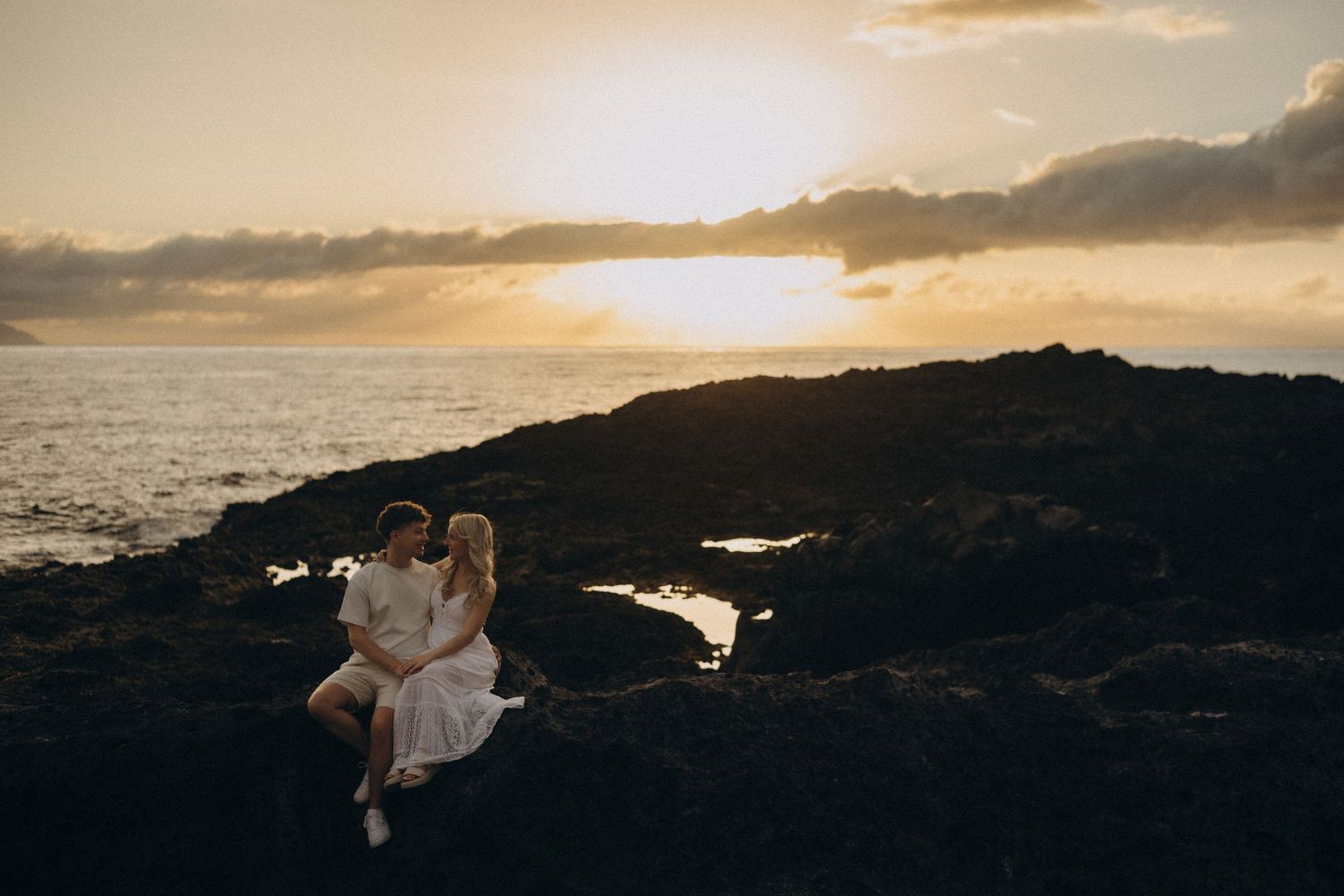 Couple sitting on volcanic rocks at sunset by the ocean, sharing a romantic moment with golden sky.