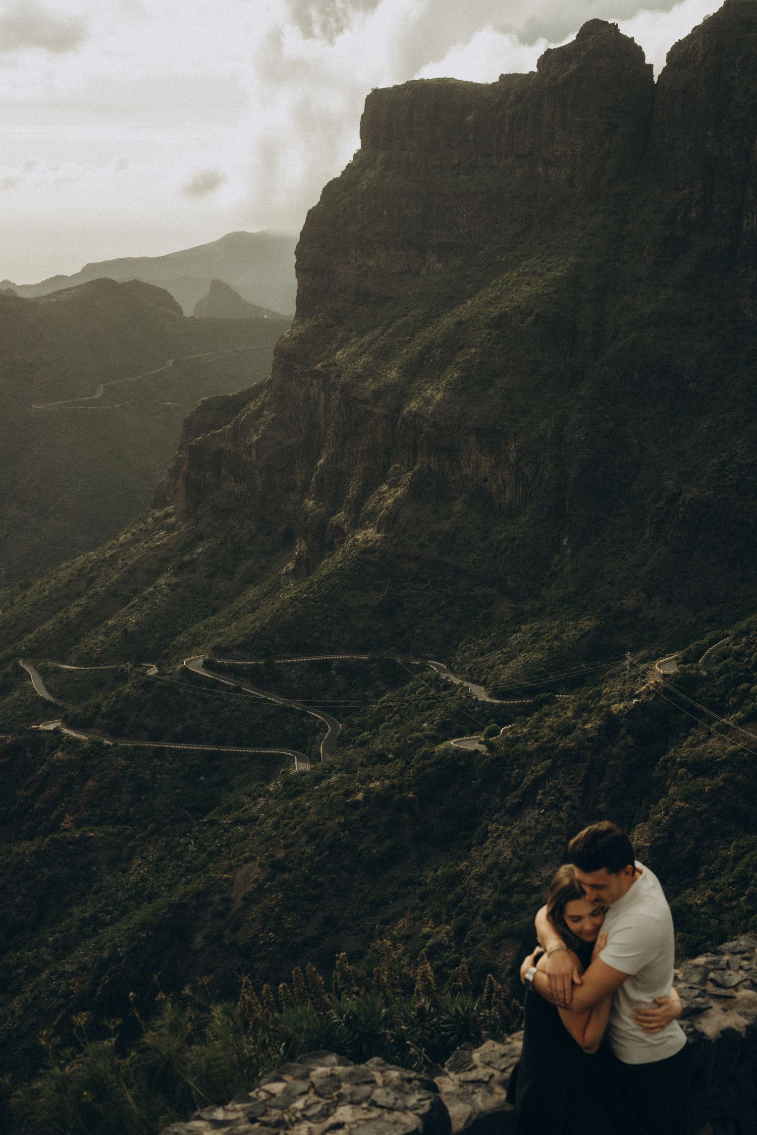 Couple embracing romantically on a rocky overlook with dramatic green mountain cliffs and winding road in background.
