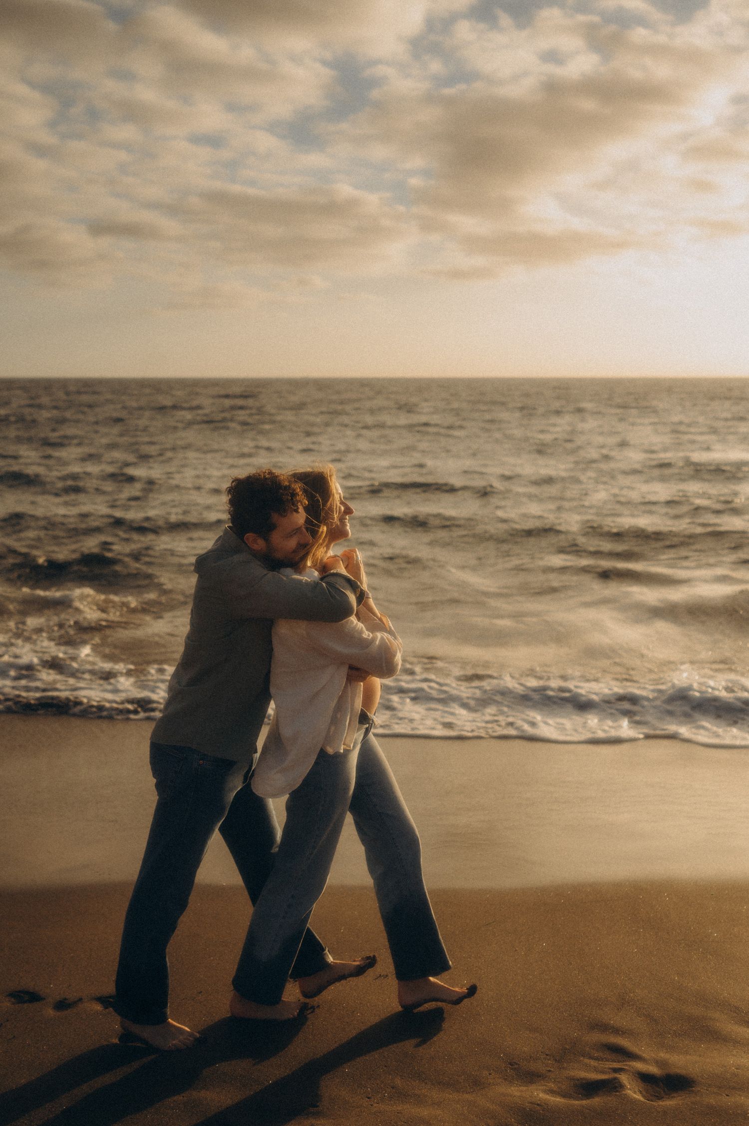 Couple embracing barefoot on sandy beach at sunset with ocean waves in the background.