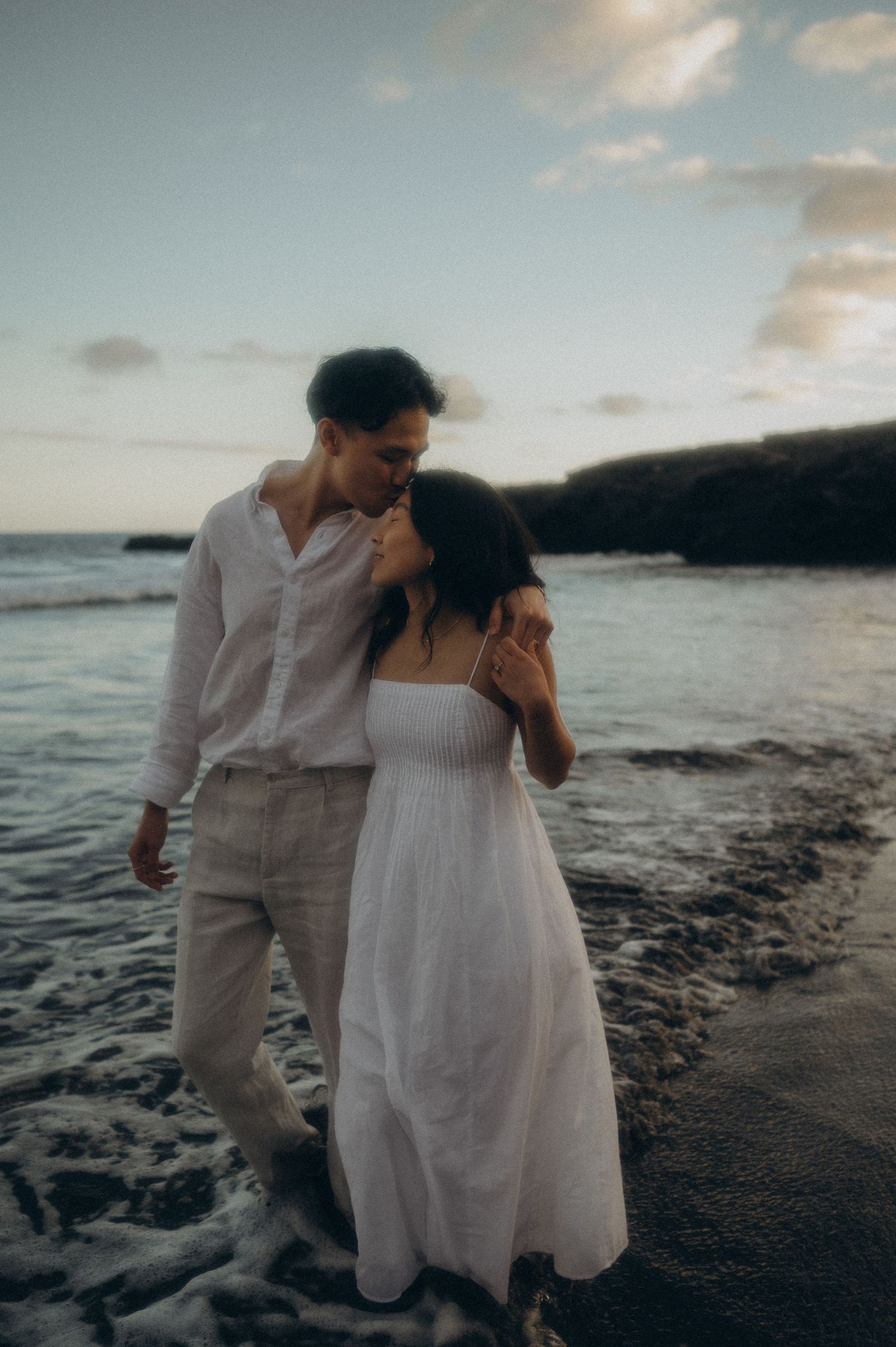 A couple in white attire shares an intimate moment on a black sand beach at sunset with ocean waves in the background in Tenerife.
