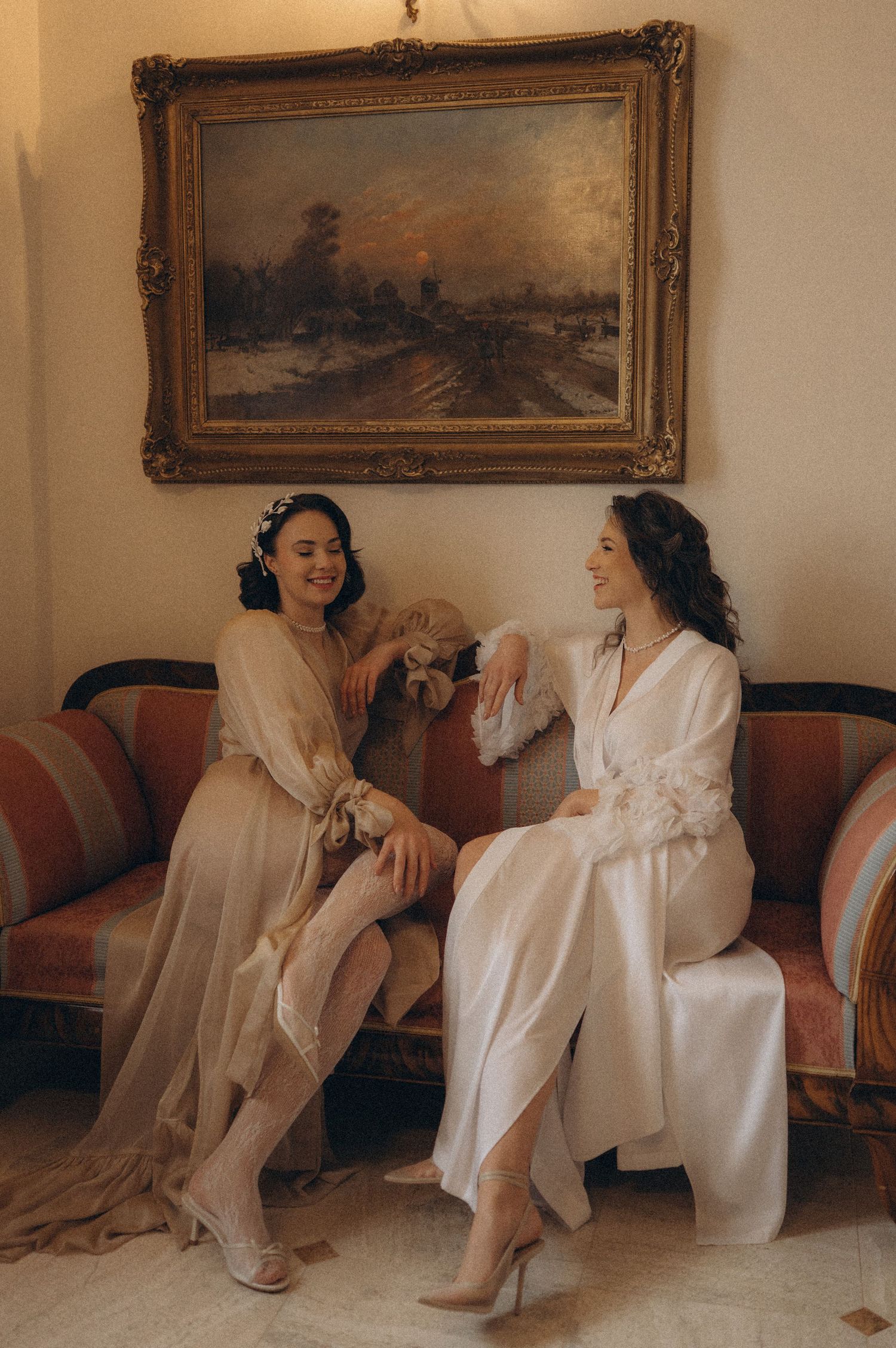 Two brides seated on a vintage-style sofa in a castle-inspired wedding editorial setting beneath a grand painting.