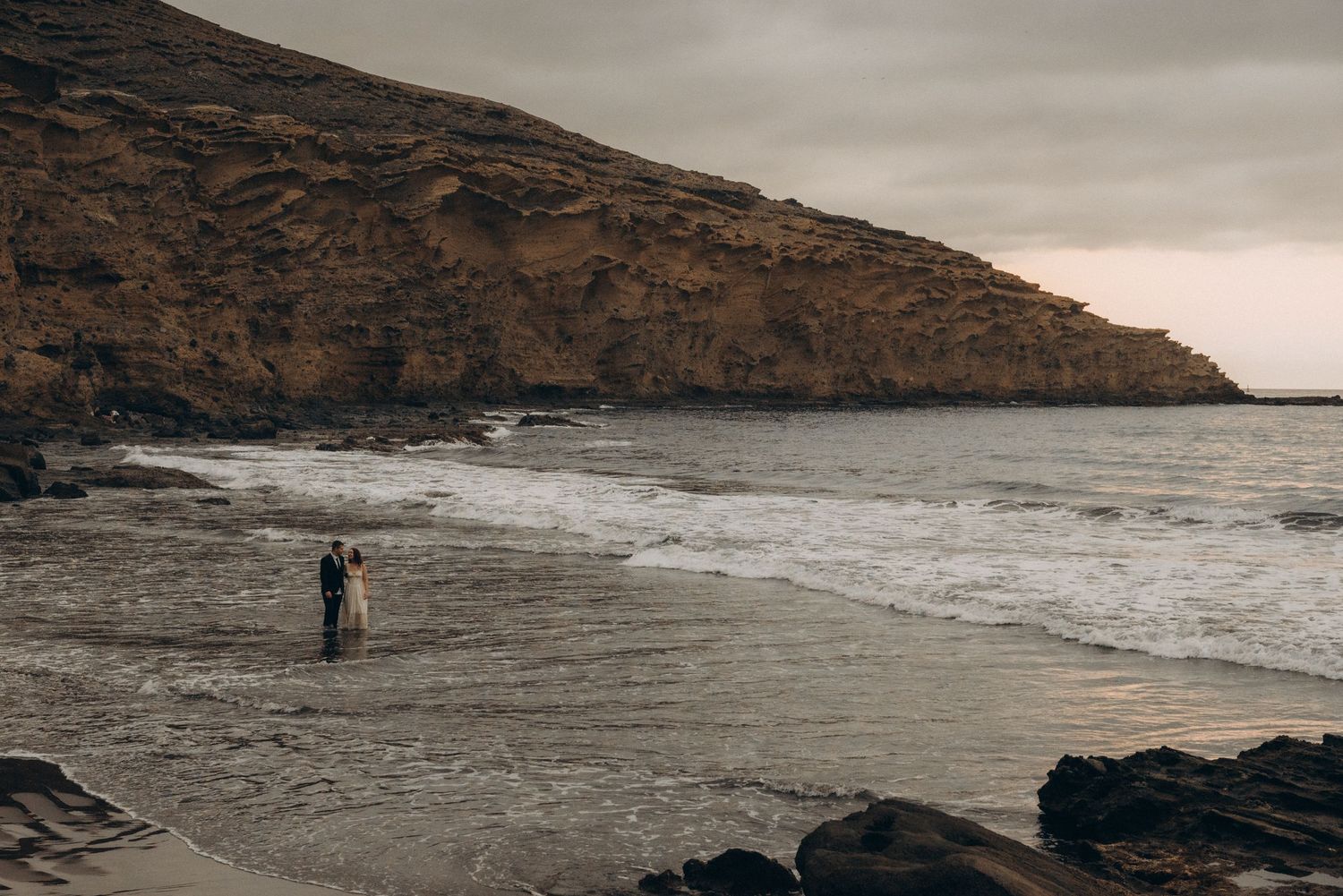 A lone figure stands on a dramatic rocky beach with crashing waves and a rugged brown hillside under overcast skies.