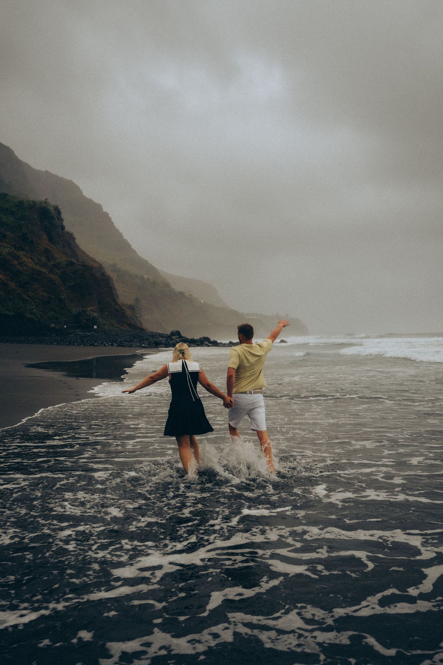 Couple holding hands running through black sand beach waves with misty mountains and overcast sky.