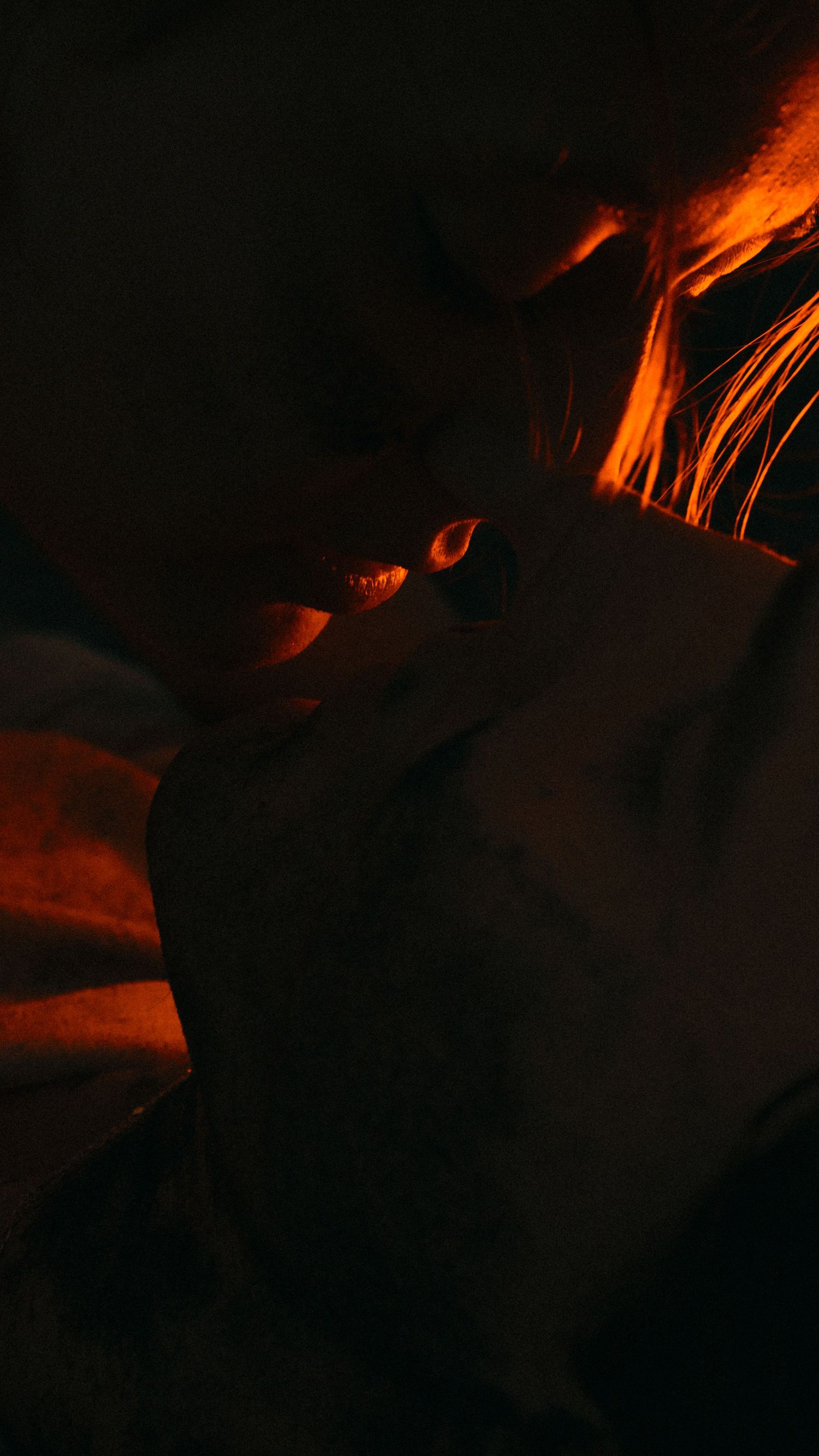Dark moody portrait with warm orange light illuminating a persons face and hair in dramatic shadows.