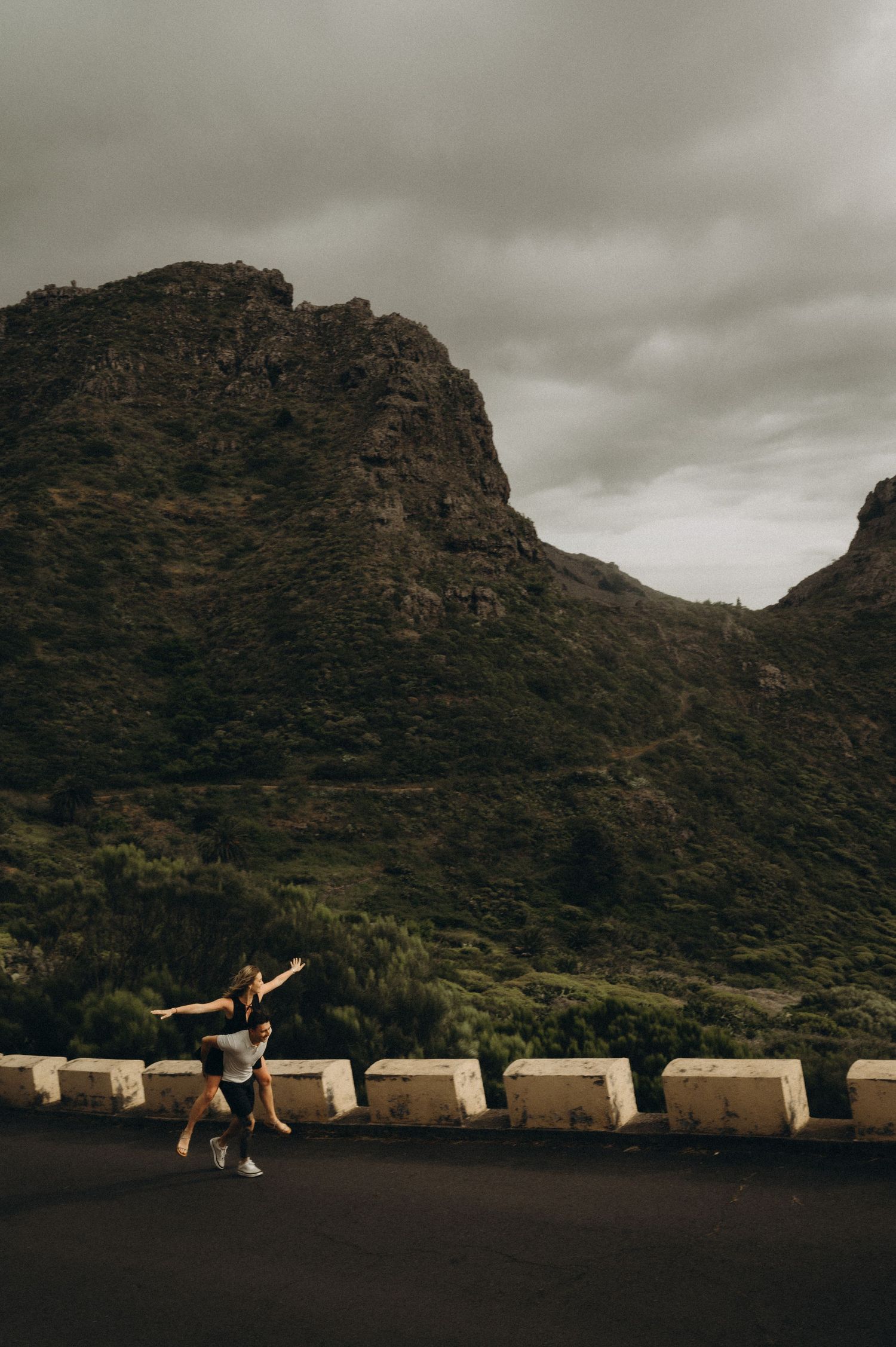 Two people dancing on a road with dramatic green mountains and cloudy sky in the background.