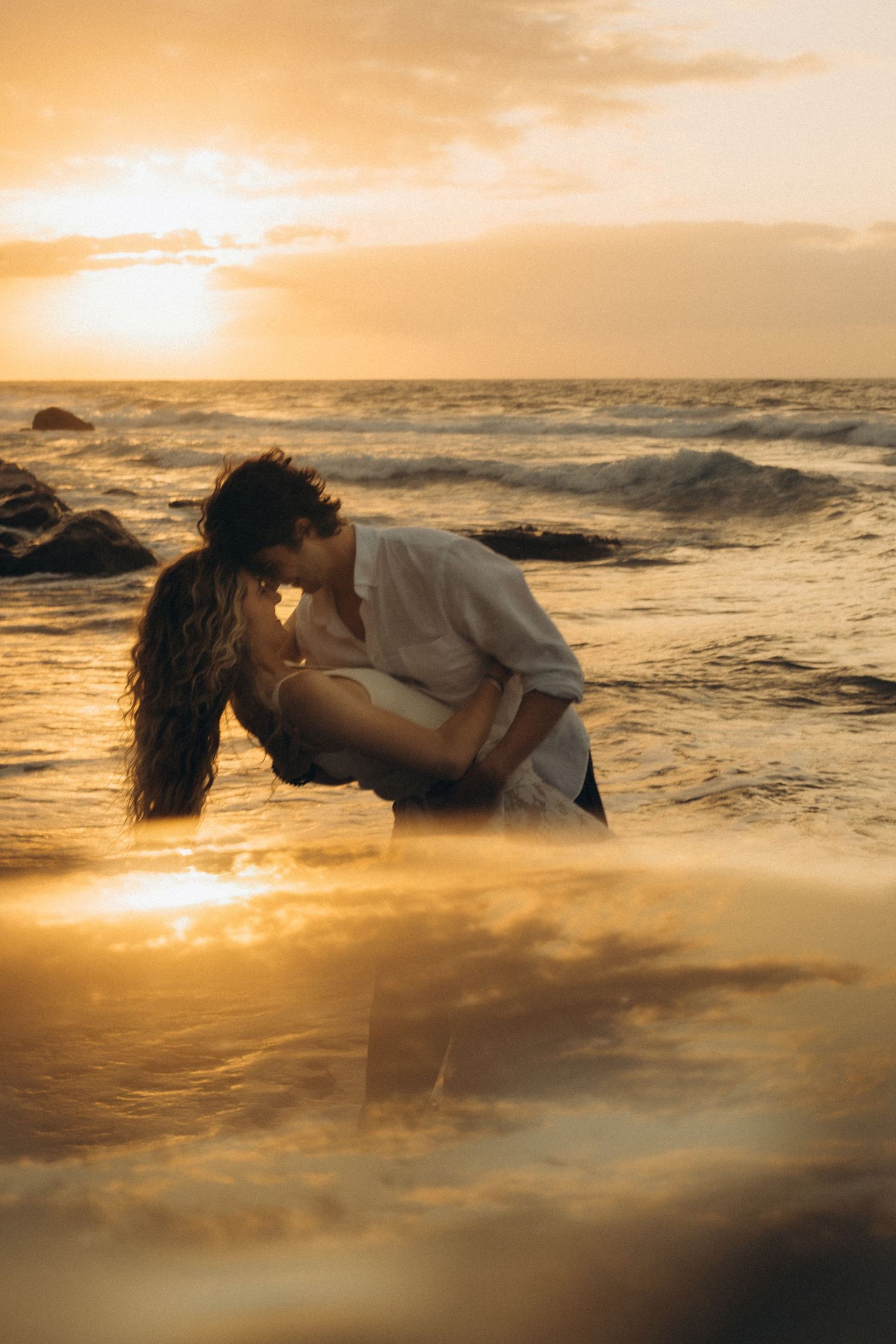 Couple sharing a romantic kiss on the beach at golden sunset, waves crashing behind them.