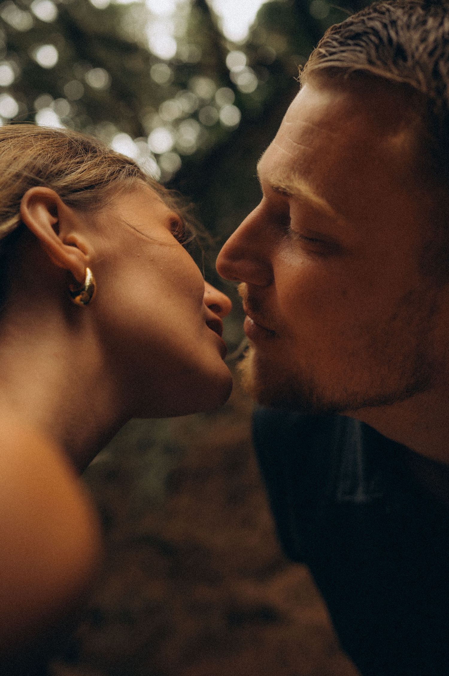A couple sharing a romantic kiss outdoors in a forest setting, with soft bokeh background lighting.