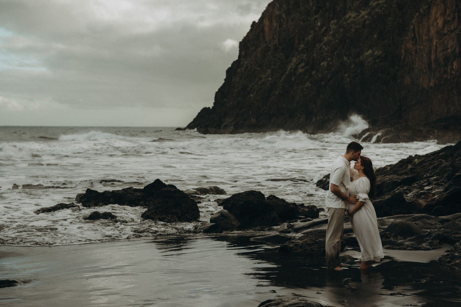 Couple embracing on rocky beach with dramatic ocean waves and cliffs under stormy skies.