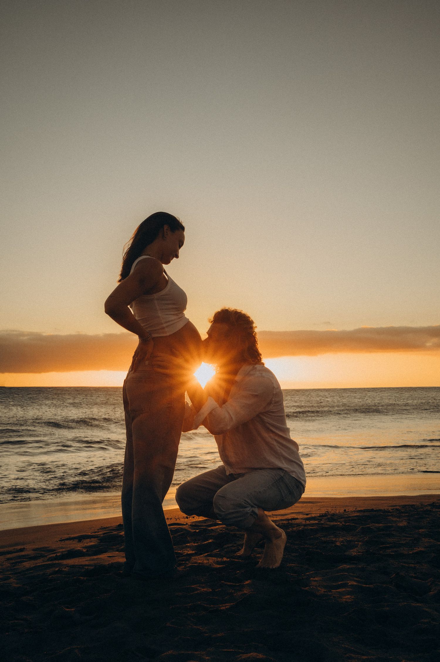 Couple sharing a tender maternity moment on a beach at golden sunset, silhouetted against the glowing horizon.