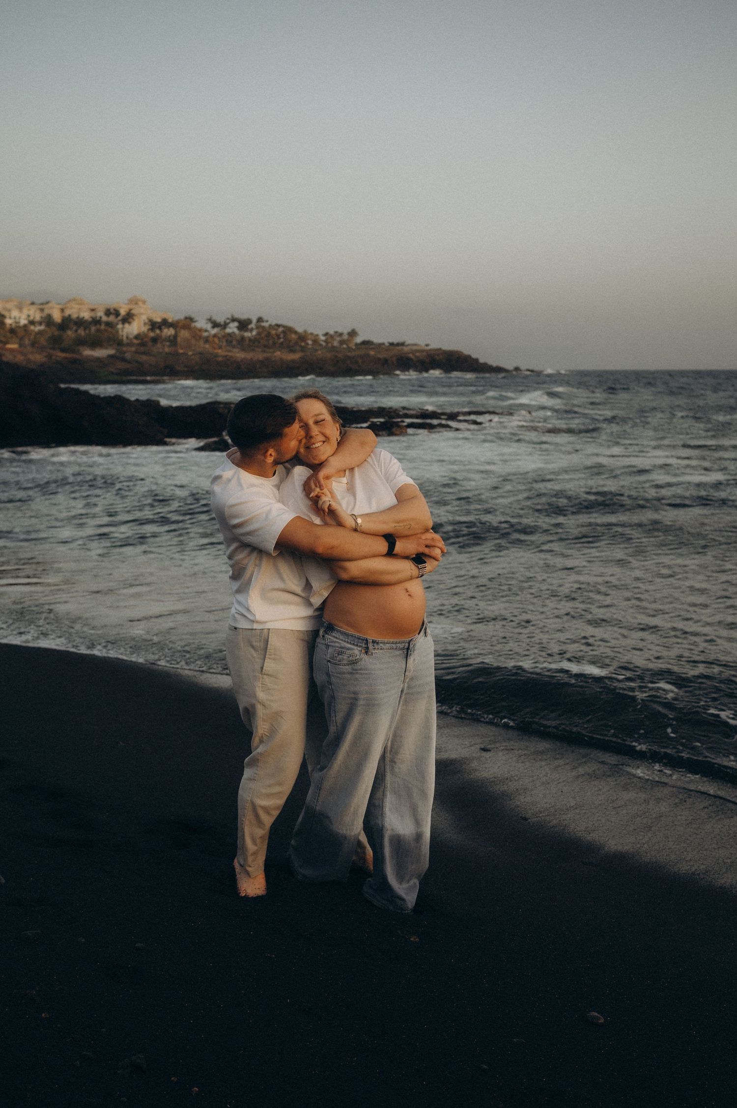 Couple embracing on black sand beach at sunset, pregnant woman in white top and jeans, waves and rocky coastline behind them.