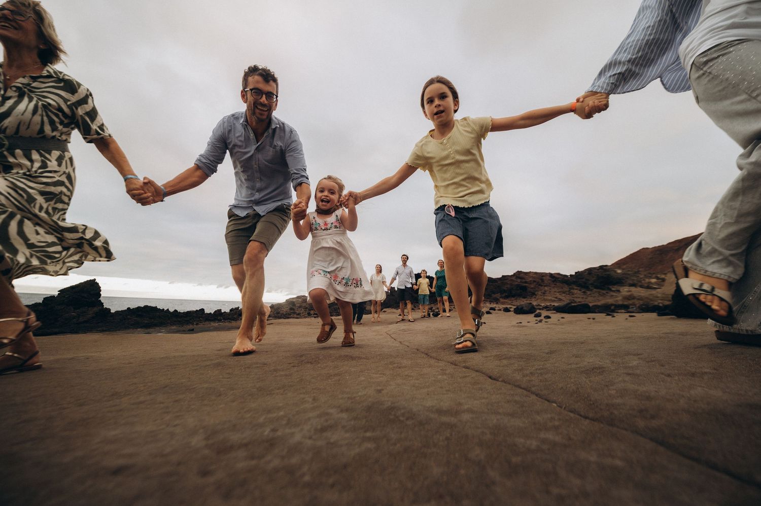 Playful family running together toward camera on wide lens along rocky coastline, Tenerife