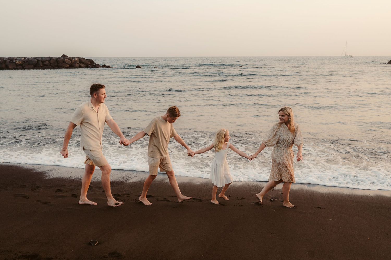 Mother leading family holding hands along black sand beach by ocean at sunset, Playa del Duque, Tenerife