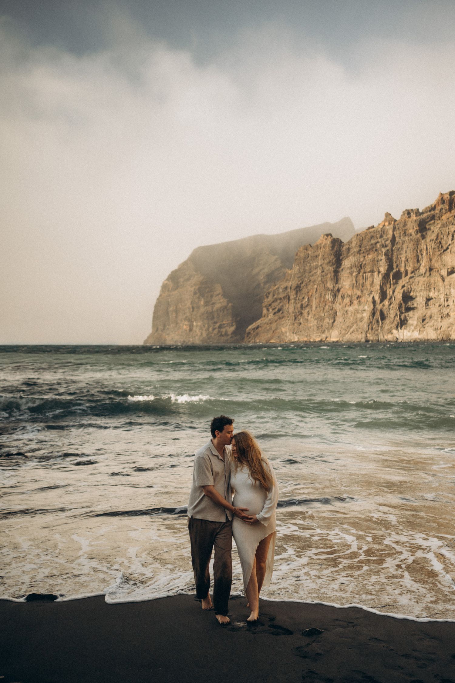 Couple embracing on black sand beach with dramatic cliffs and ocean waves in the background.