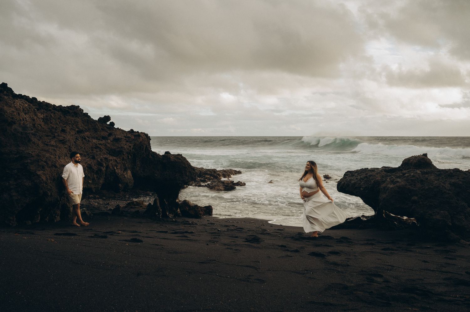 Couple in white outfits on a black sand beach with dramatic rocky coastline and crashing waves under cloudy skies.