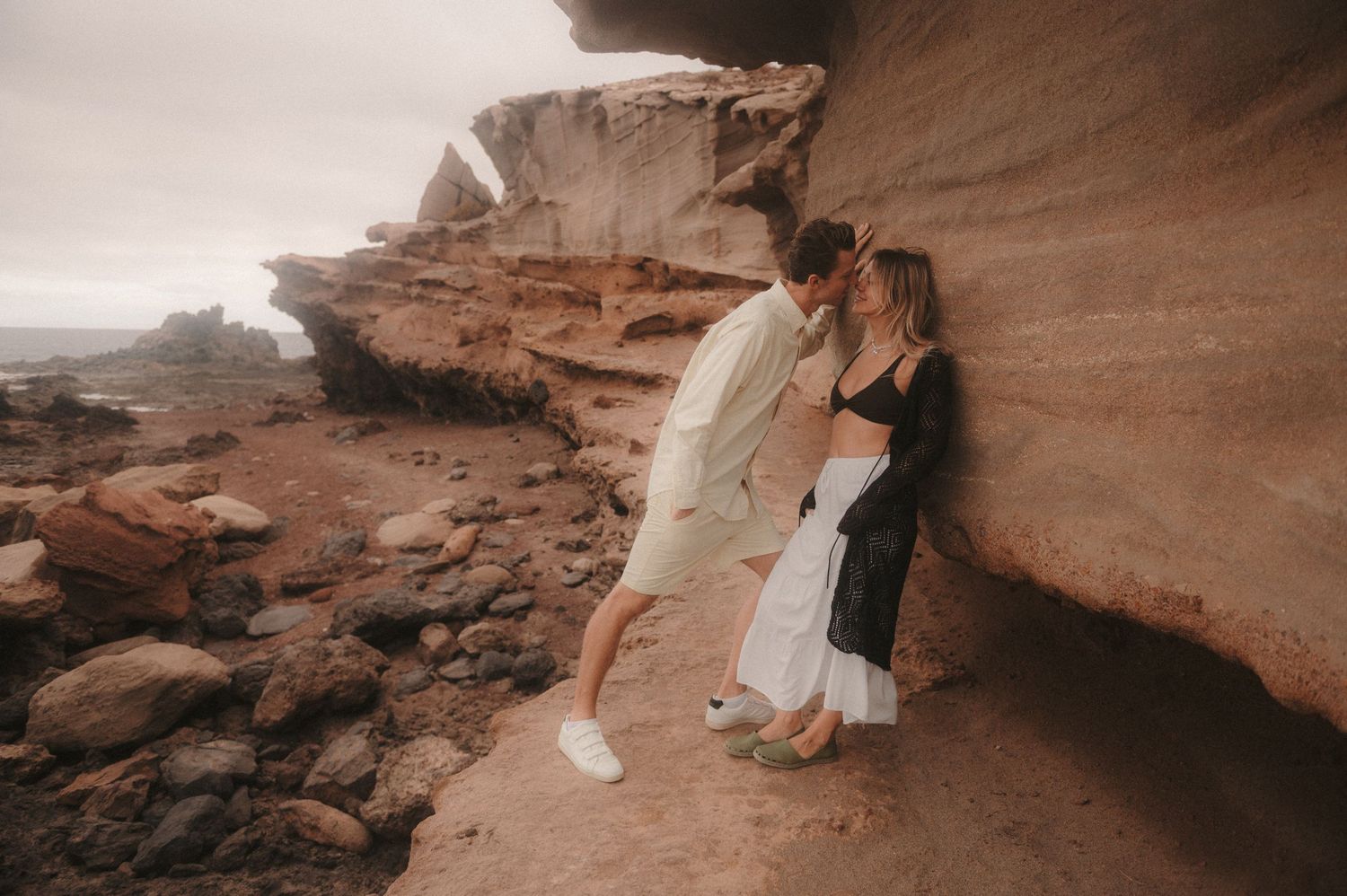 Couple by a colorful cliff, she leaning with her back against it while he leans in to kiss her, romantic photoshoot moment in Tenerife