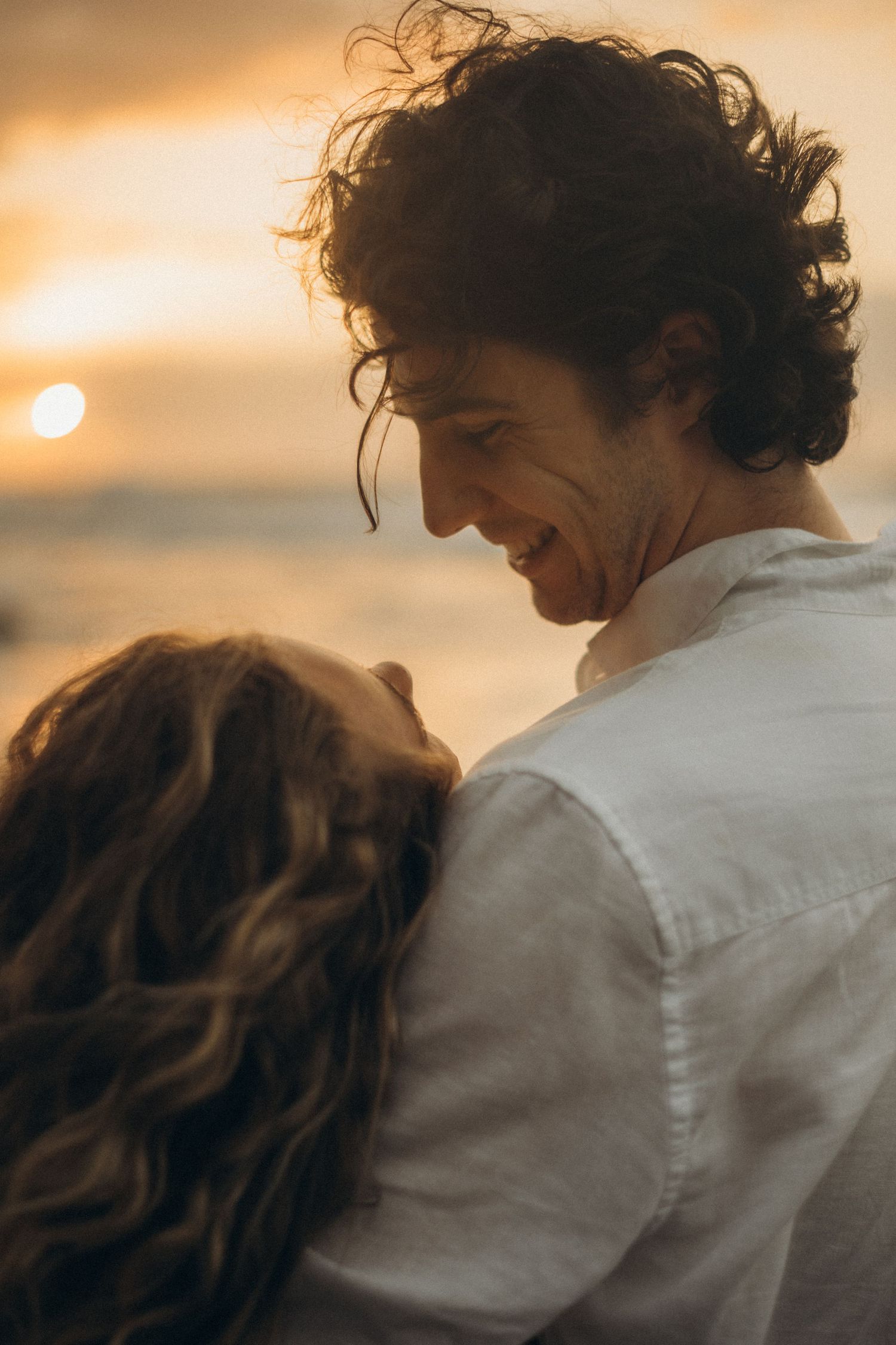 A joyful couple embraces on a beach at sunset, the man laughing warmly as golden light glows behind them.