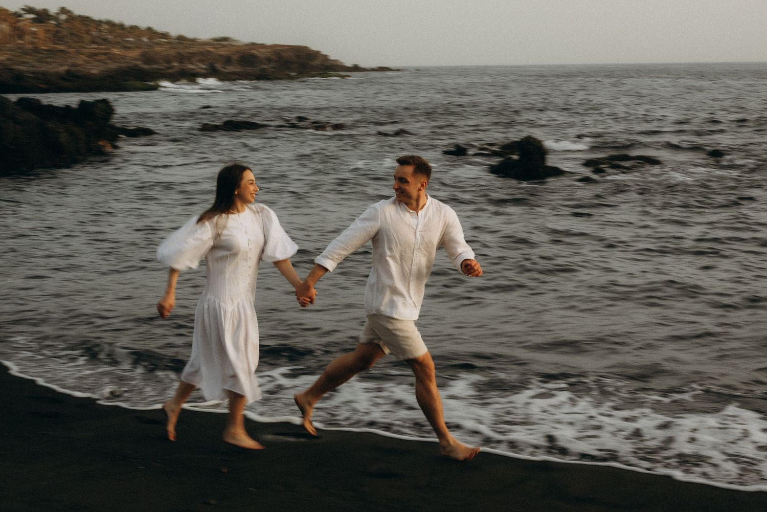 Couple in white outfits holding hands and running on a black sand beach with ocean waves and rocks.