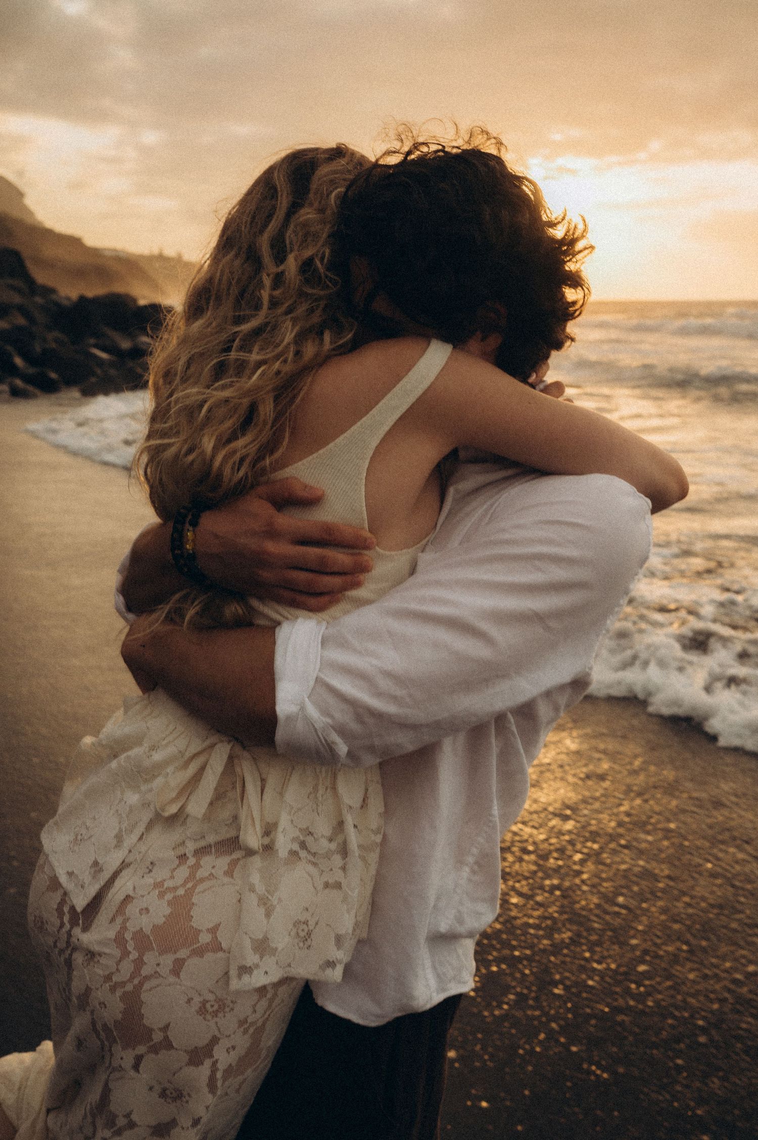 Couple embracing romantically on a sandy beach at golden sunset with ocean waves in the background.