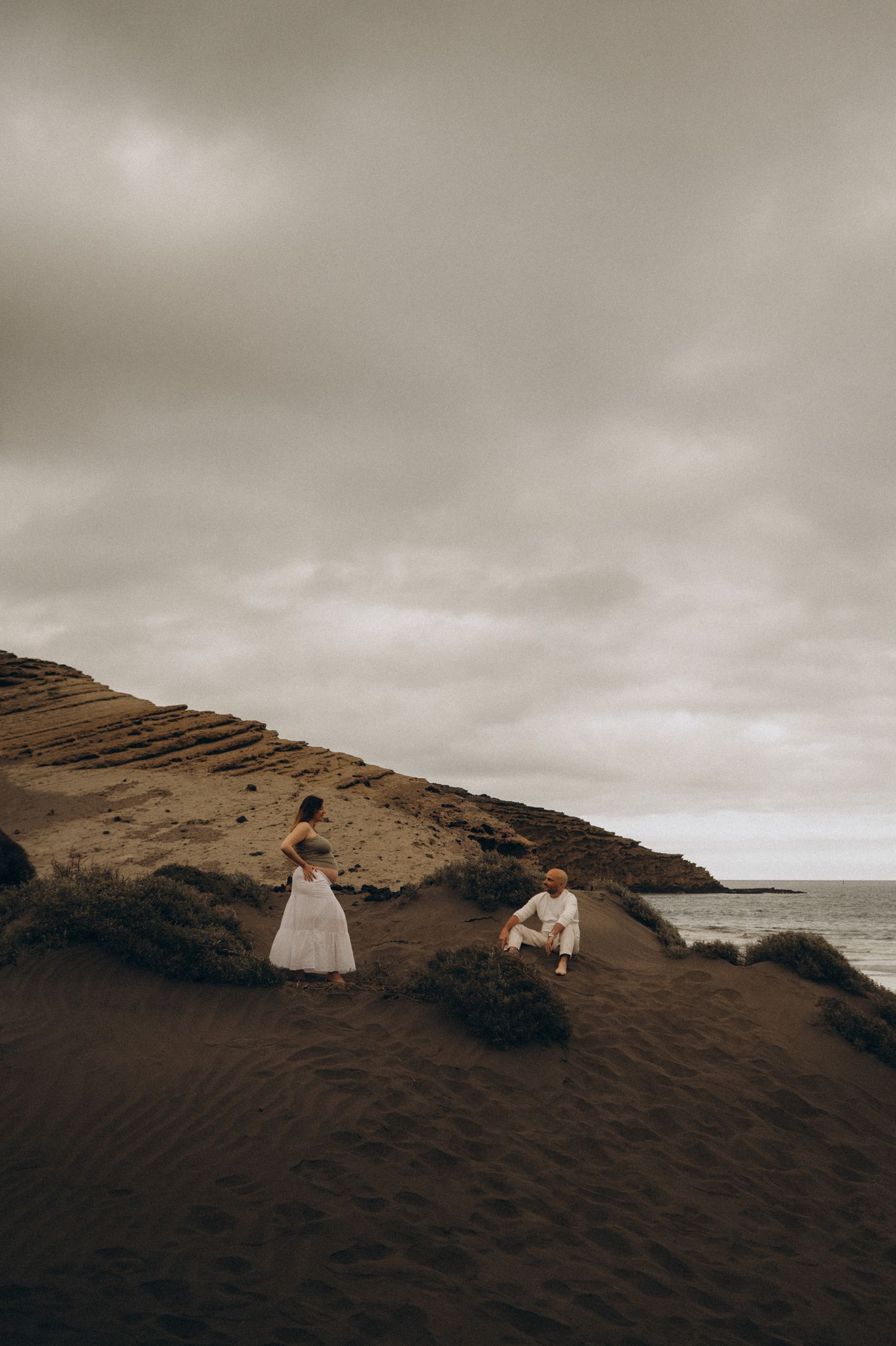 Couple in white attire on dark volcanic sand beach with dramatic cloudy sky and ocean backdrop.