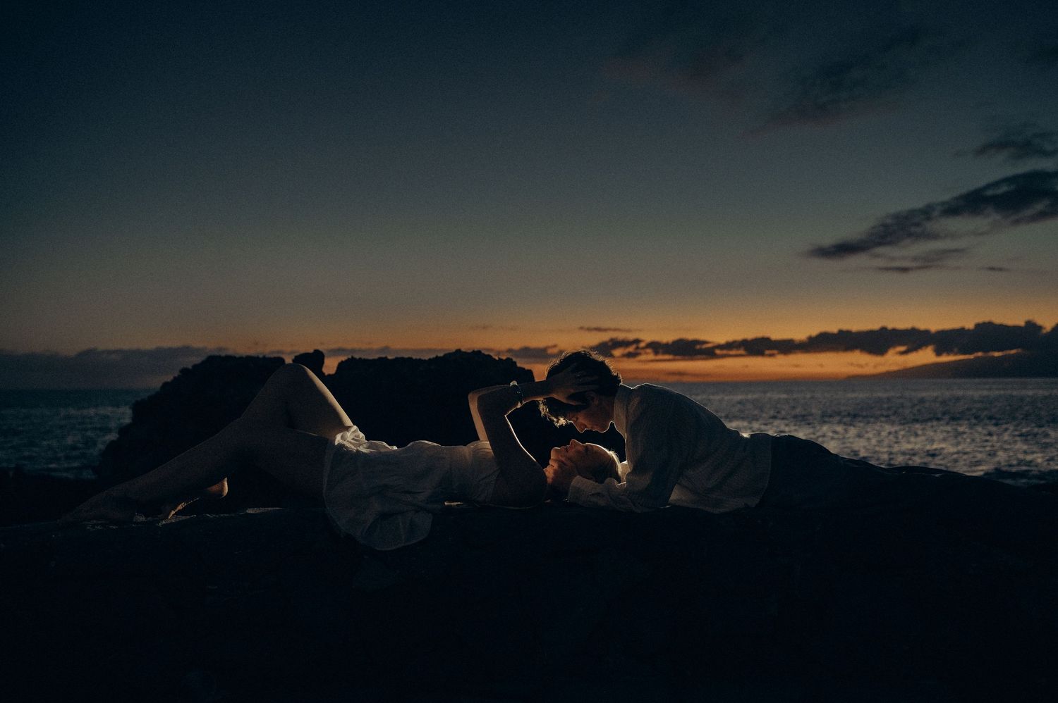 Couple sharing a romantic kiss on rocky beach at sunset with ocean waves and colorful sky in background.