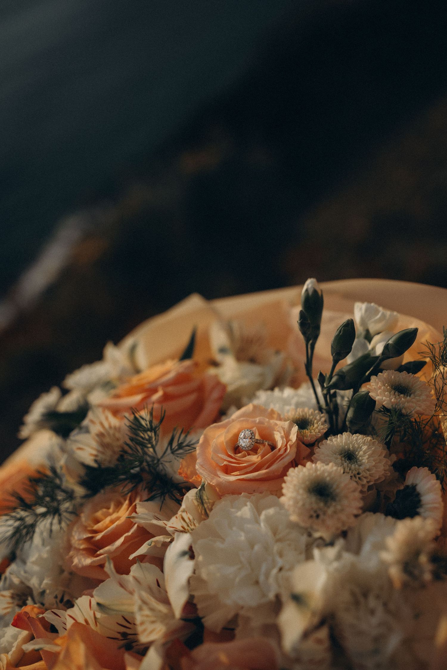 A romantic bouquet of peach roses and white flowers with a diamond engagement ring nestled among the blooms.