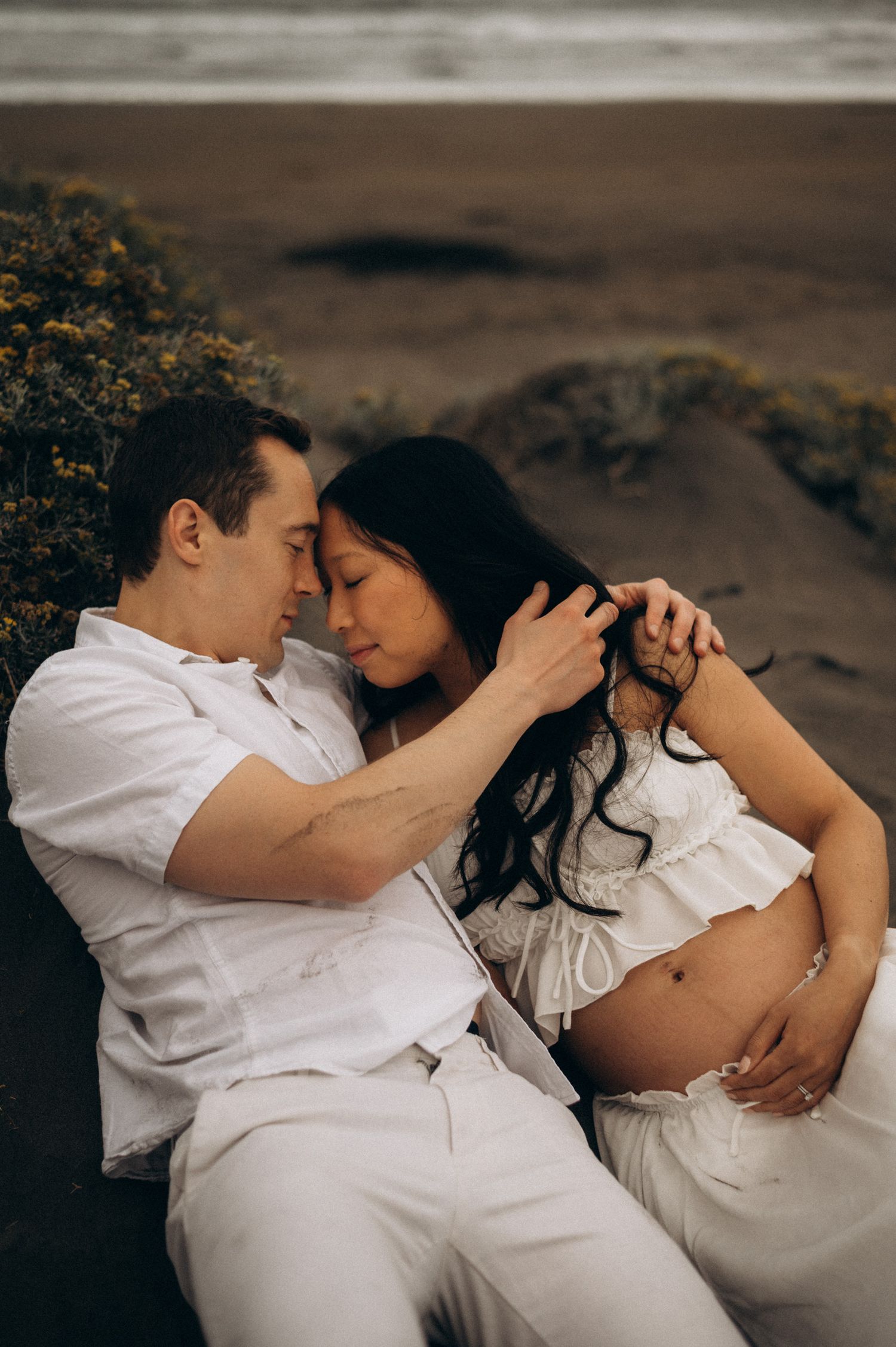 Couple in white outfits sharing tender moment on beach, pregnant woman cradling baby bump while partner gently touches her hair.