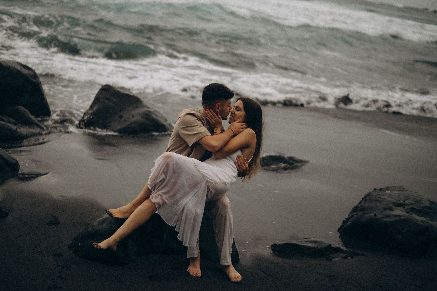Romantic couple kissing on a dark volcanic beach with dramatic ocean waves and rocks in the background.